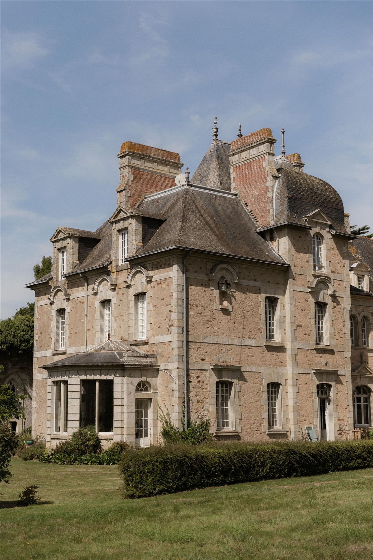 An exterior wide shot of a French château photographed from the grounds, with no people visible. The building is constructed from pale sandy-pink granite stone with white-trimmed arched and rectangular windows fitted with white shutters. The architecture features multiple steep slate-grey pitched roofs, a central conical tower, decorative stone finials, brick chimney stacks, and a ground-floor glass conservatory or orangerie with a pitched glass roof. A trimmed low hedge runs along the base of the building, and a manicured lawn extends into the foreground. Potential venue feature image.