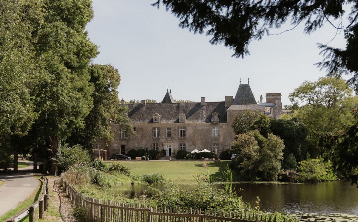 A wide exterior shot of a large French château with pale stone walls, steep slate-grey mansard roofing, dormer windows, and pointed corner towers with decorative finials. The building is photographed from across a pond with lily pads in the foreground, with a wooden post-and-rail fence lining a gravel driveway to the left. A well-kept lawn stretches between the pond and the château's entrance, where two white parasols and parked vehicles are visible near the front doors. The grounds are framed by mature trees on both sides. Potential venue feature image.