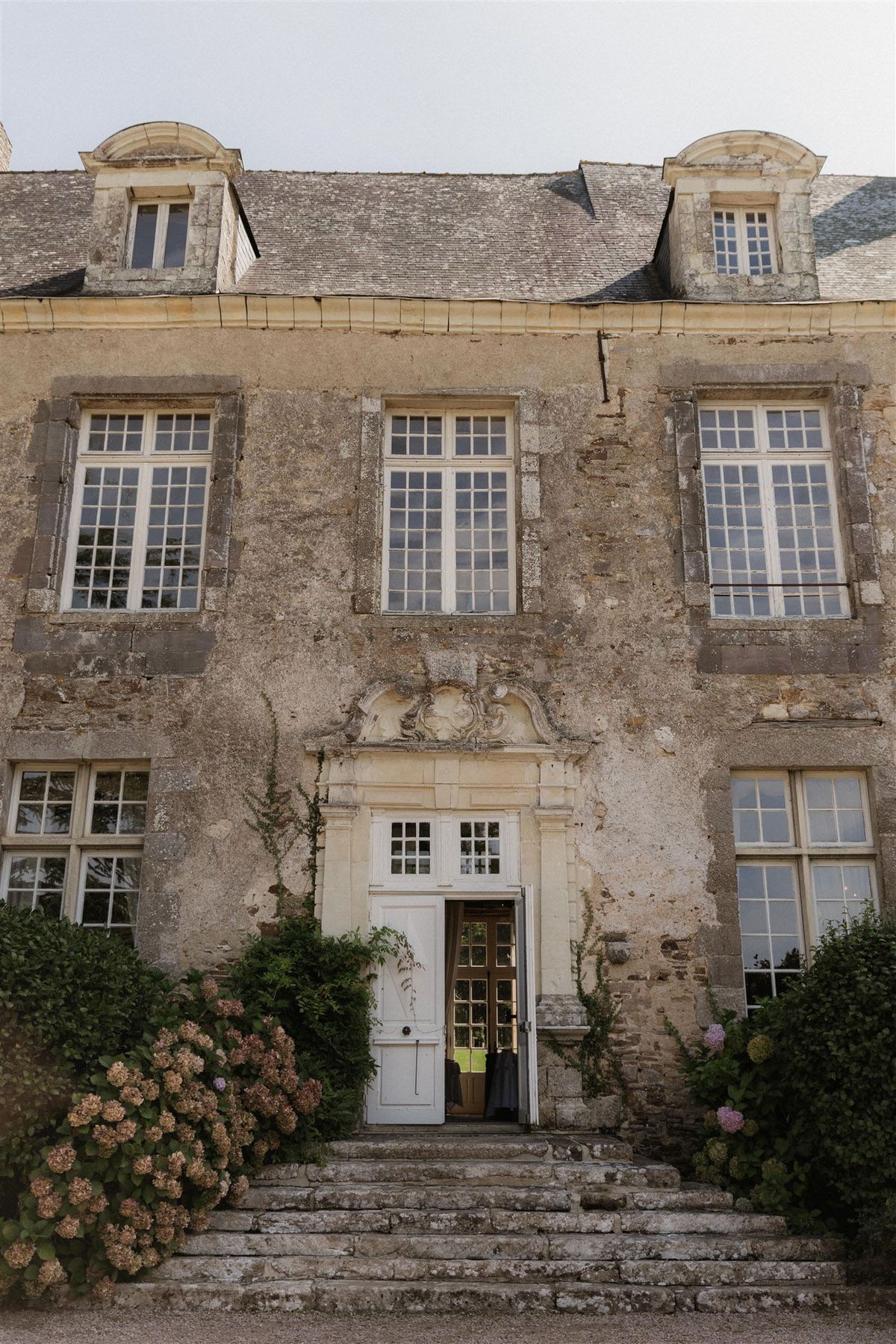 A straight-on exterior shot of a French château or manor house, showing the full front facade in a portrait orientation. The building features aged limestone or granite stone walls, white-painted multi-pane windows across three floors, and two dormer windows on the upper mansard-style roof. The central entrance has an ornate carved stone pediment above a white painted double door, which stands open revealing an interior glimpse. Stone steps lead up to the entrance, flanked by climbing vines on either side of the doorway. Blush-pink and dusty mauve hydrangea bushes are visible at the lower left and right corners of the facade, adding decorative color against the weathered stone. No people are visible in the frame. Potential venue feature image.