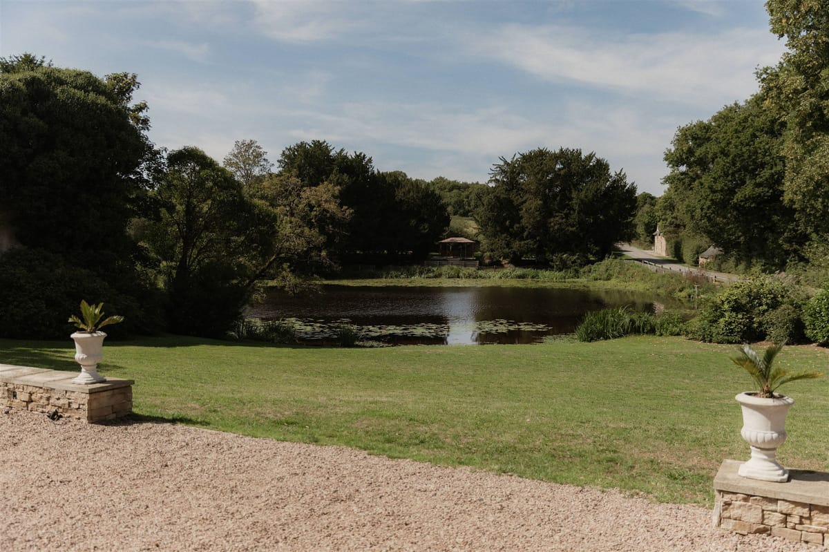 A wide shot of a French estate's grounds showing a ornamental pond with lily pads in the middle distance, framed by a manicured lawn. Two white classical stone urns planted with tropical foliage are positioned on low stone plinths in the foreground, flanking a gravel terrace or driveway. A small wooden gazebo or pavilion structure is visible on the far bank of the pond. No people are present in the image. Potential venue feature image.