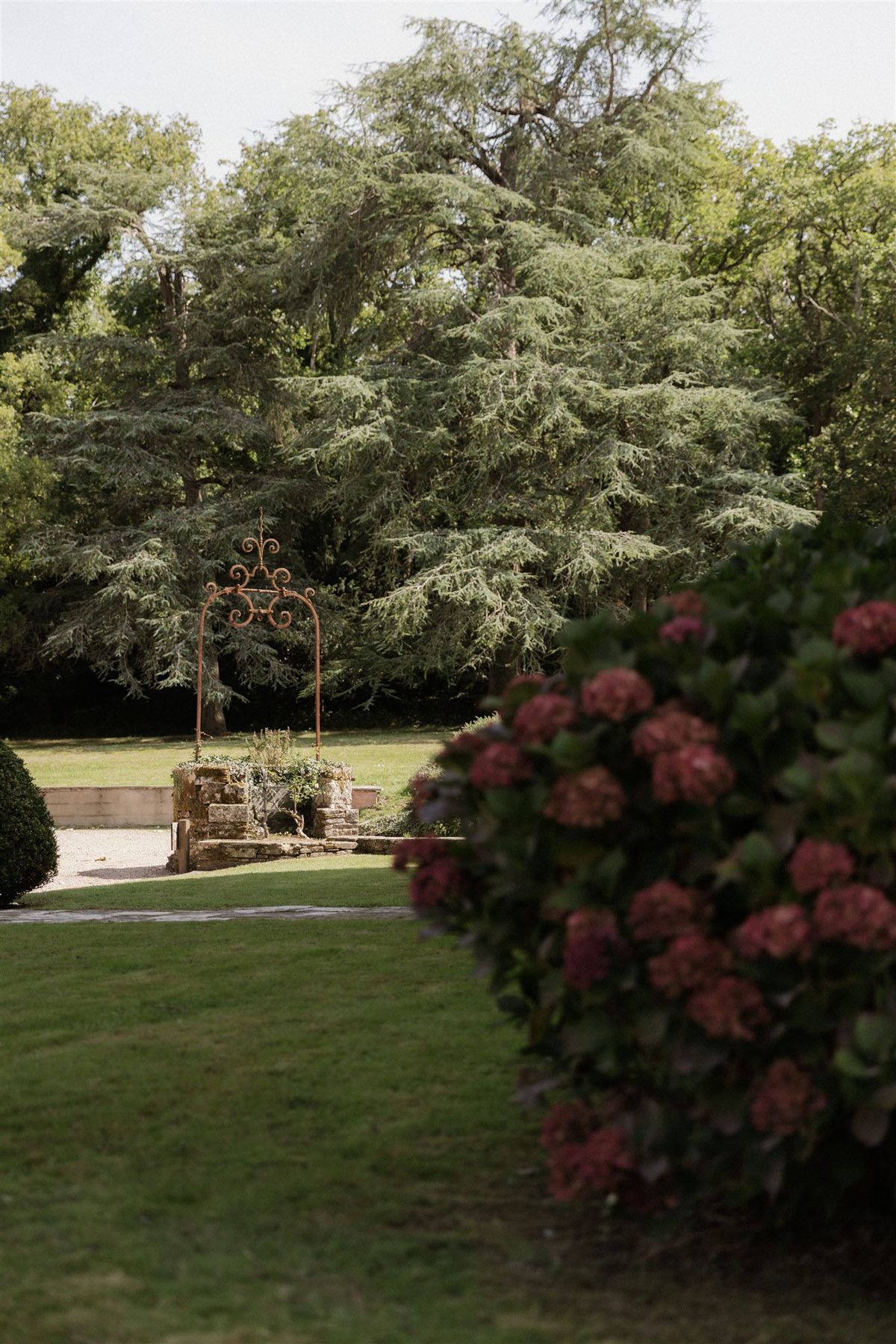 A wide shot of formal garden grounds featuring a rusty wrought-iron decorative arch with scrollwork detail, positioned above a low stone well or planter structure at the center of a gravel pathway. The foreground right is occupied by a large hydrangea bush with deep pink-to-mauve blooms, rendered slightly out of focus. Manicured lawn stretches across the mid-ground, with clipped topiary spheres visible to the left. No people are present in the image. Potential venue feature image.