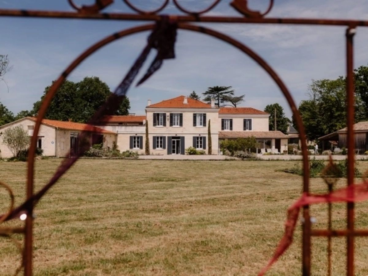 A wide exterior shot of a French country estate viewed through a rusty wrought-iron decorative arch in the foreground, which has red ribbon tied to it. The main house is a two-storey stone building with a terracotta-tiled roof, grey shutters, and a central glass door, flanked by lower outbuildings on either side. A large open lawn extends between the arch and the house. No people are visible in the frame. Potential venue feature image.