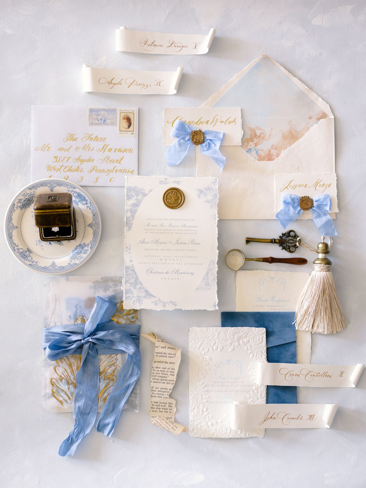 Overhead invitation suite flat lay with soft blue ribbons, gold wax seal, calligraphed envelopes and vintage ring box