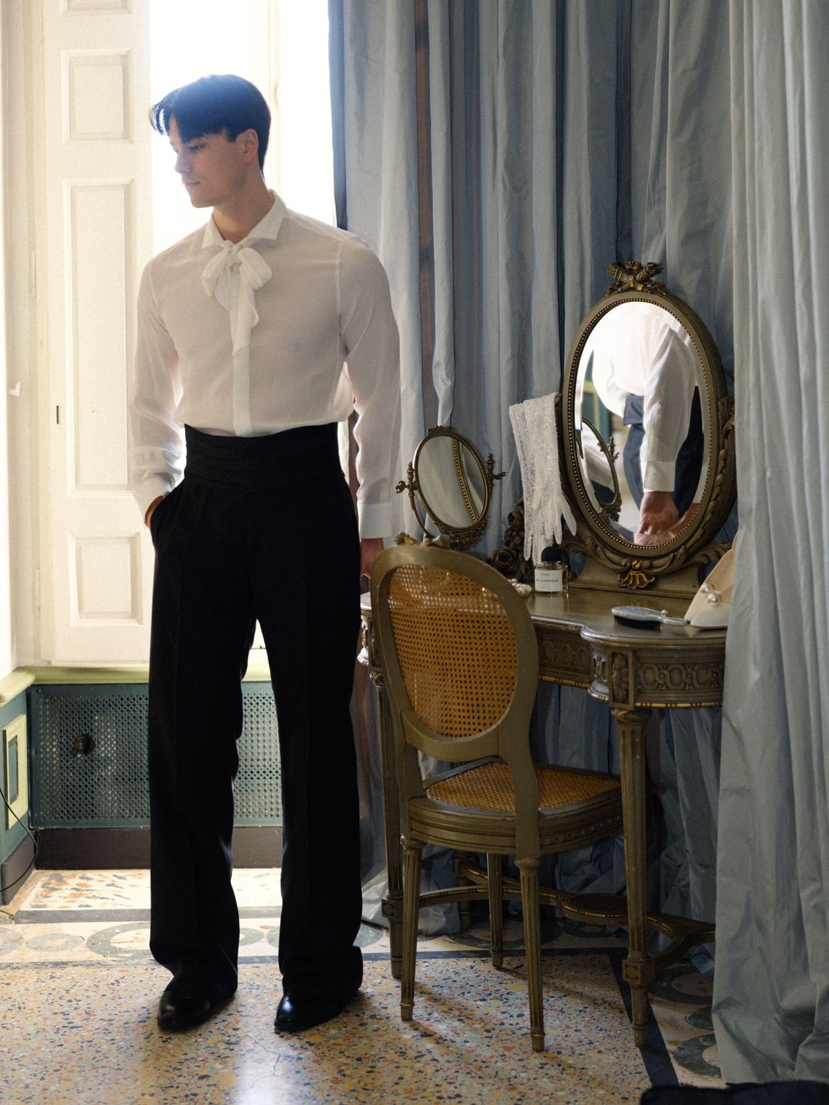 Groom dressing in white pussy-bow shirt beside antique gilt vanity in pale blue château dressing room