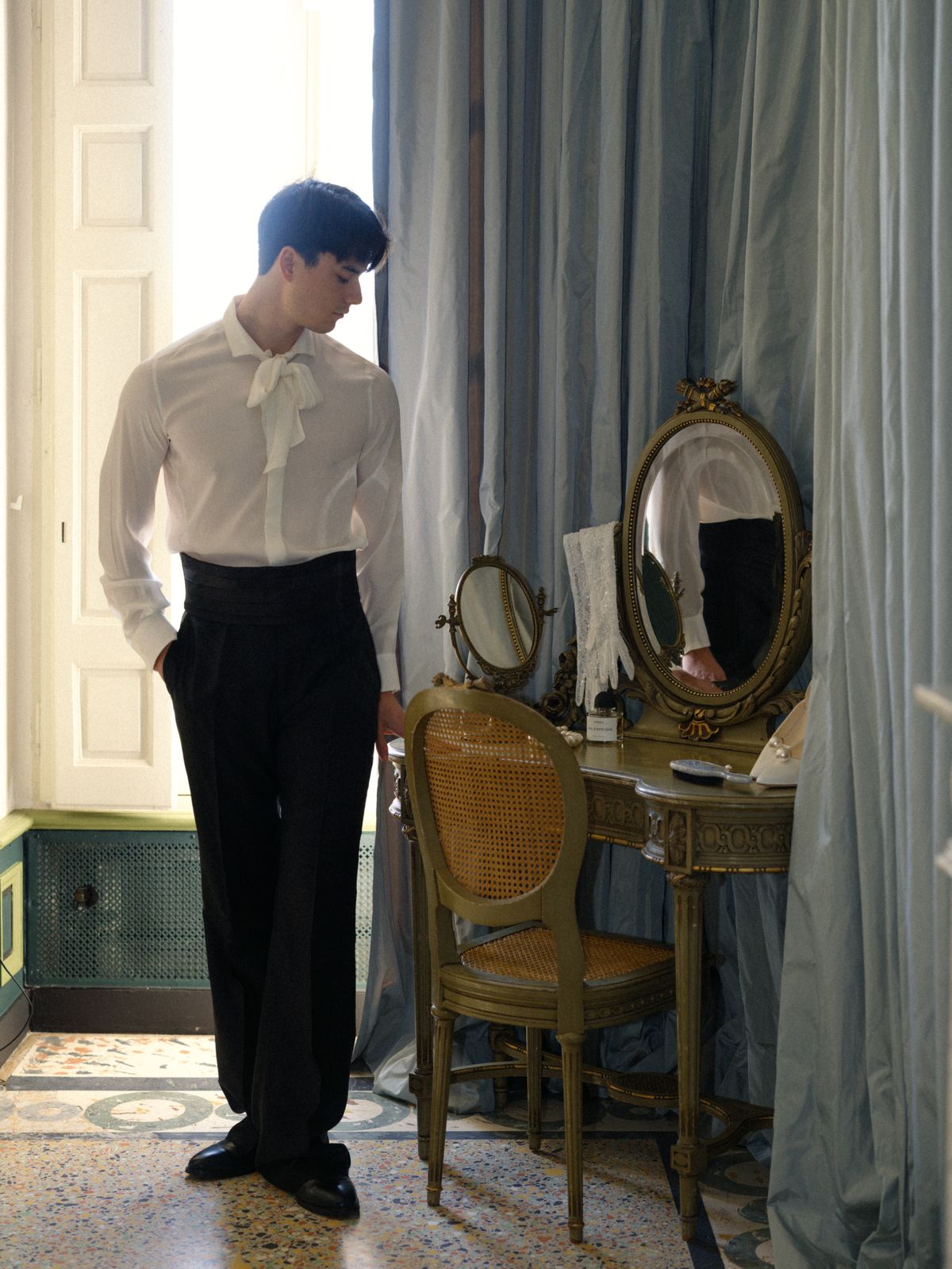 Groom in silk pussy-bow shirt beside gilt mirror and vanity in a soft blue château dressing room