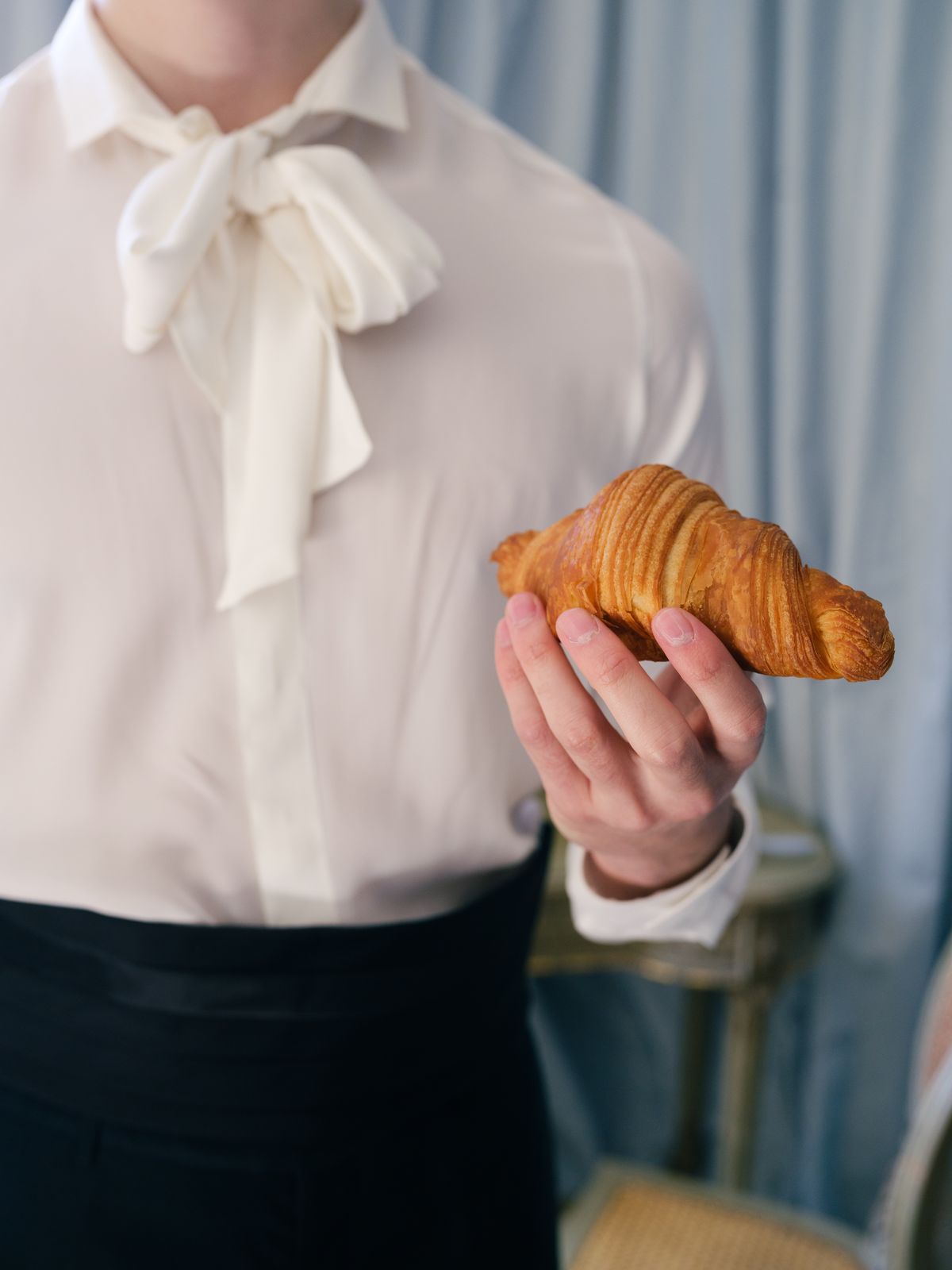 Groom in white pussy-bow shirt holding a croissant in front of pale blue château curtains