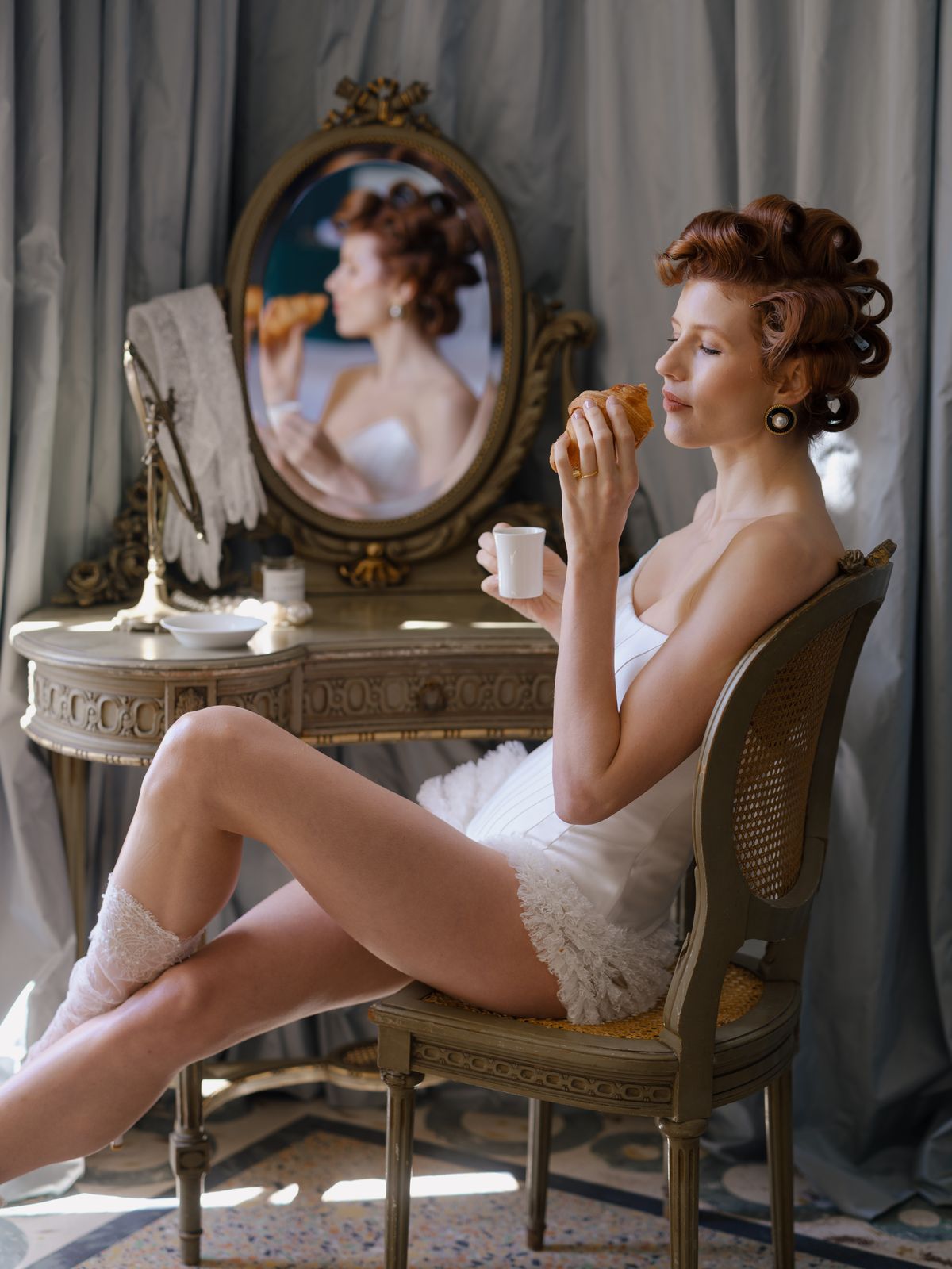 Bride reclines on caned vanity chair with cup and croissant in editorial styled getting-ready portrait