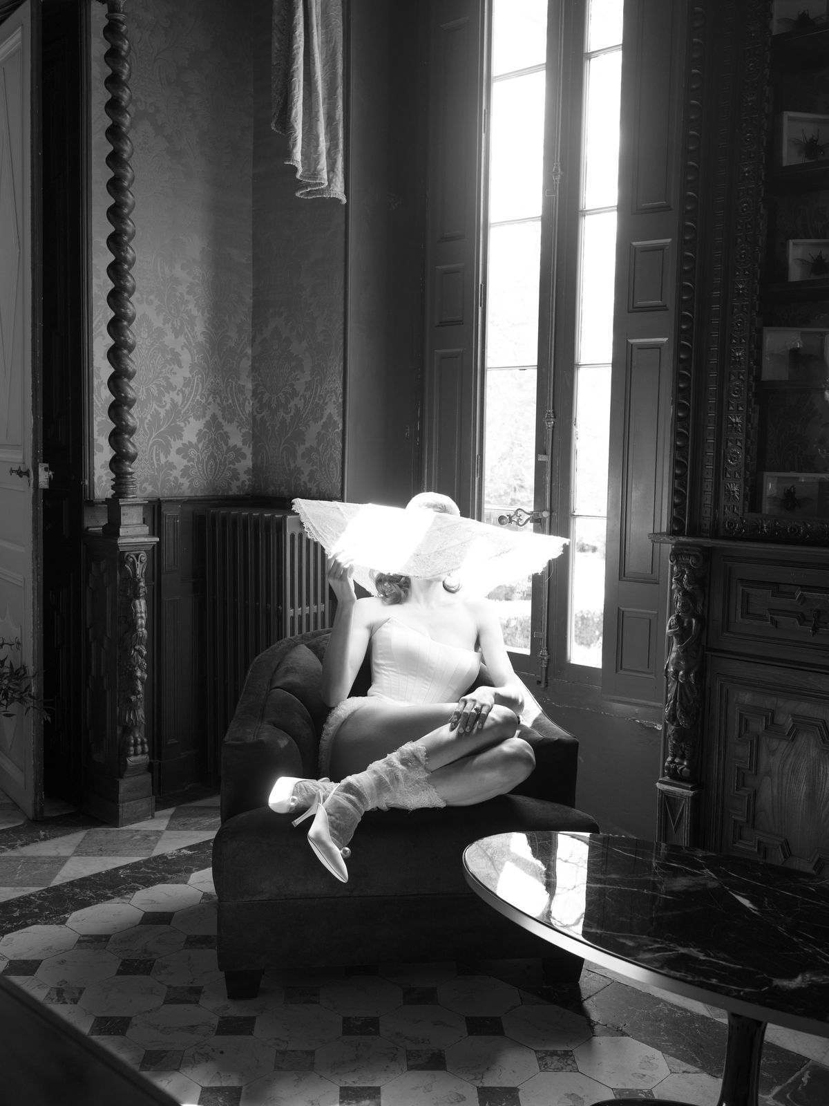 Black and white bride seated on velvet sofa in château salon with wide-brimmed lace sun hat
