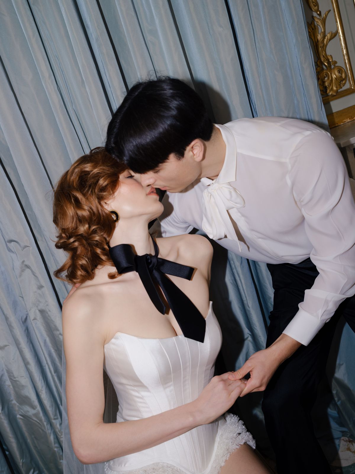 Couple kisses against pale blue silk drapes with black ribbon bow at bride's neck