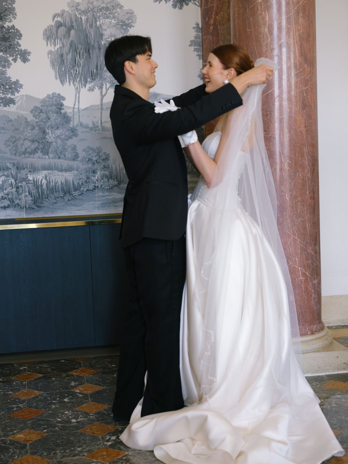 Groom lifts bride's long veil in marble columned château hall beside painted toile mural