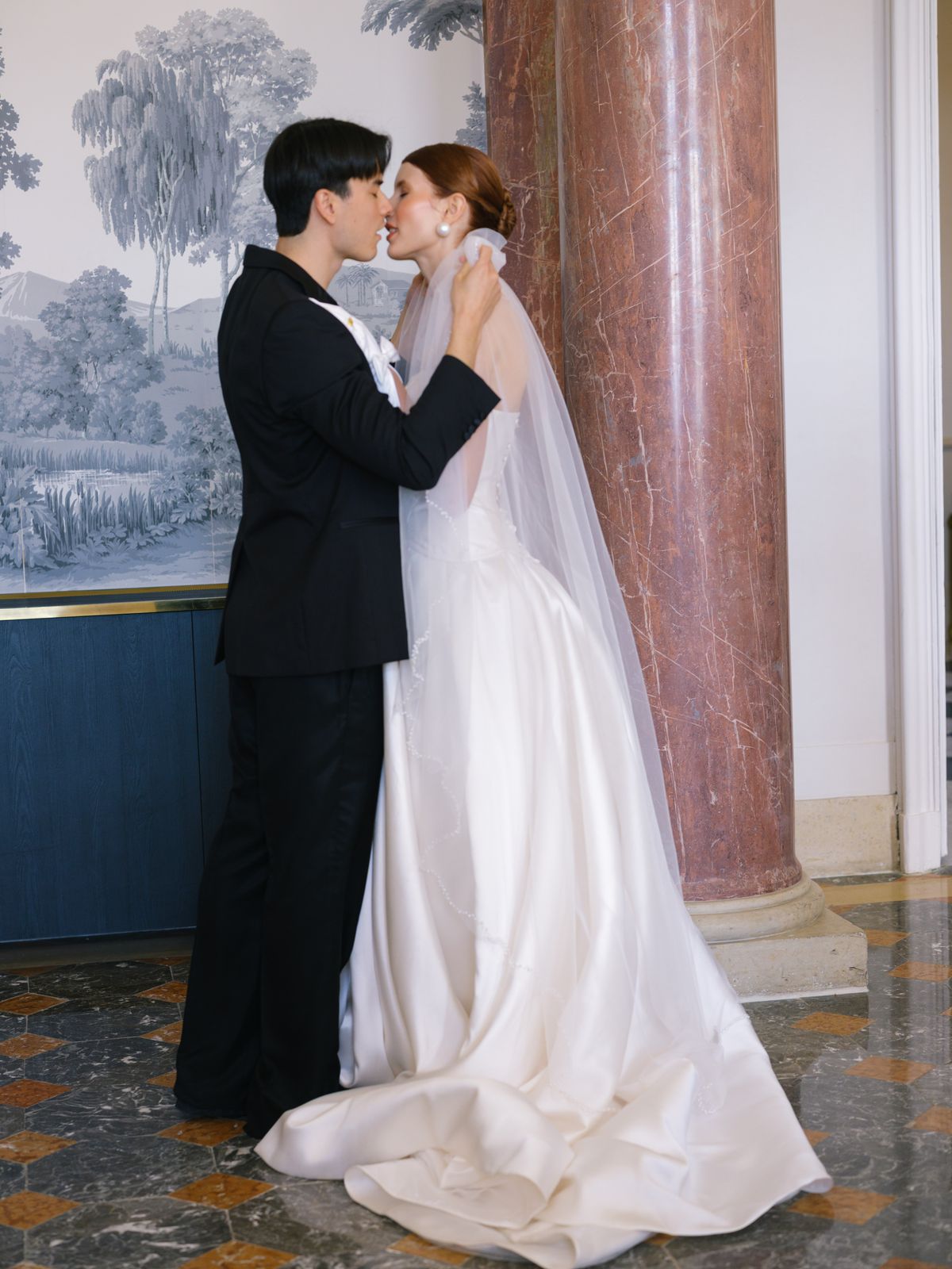 Couple kisses beside rose-marble column with bride in A-line gown and cathedral veil