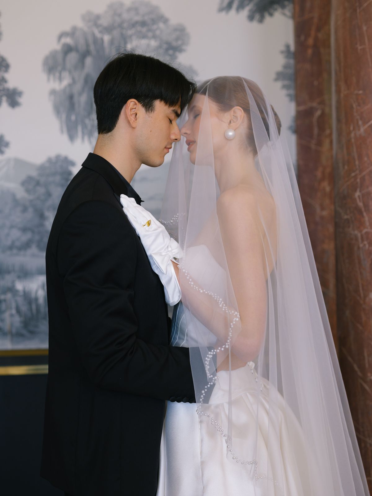 Couple faces each other under pearl-trimmed cathedral veil, bride in A-line gown and groom in tuxedo