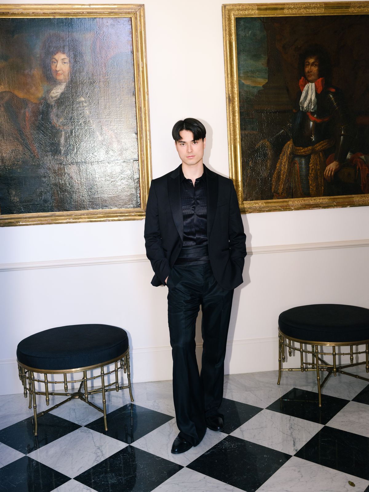 Groom in black satin tuxedo between two gilt-framed historic portraits on checkerboard marble floor
