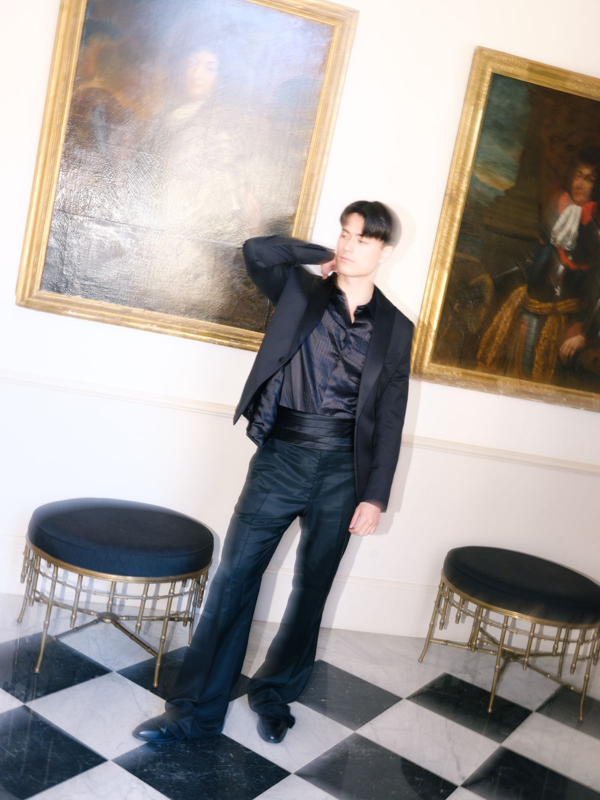 Groom poses in black satin tuxedo with hand at collar between historic gilt portraits