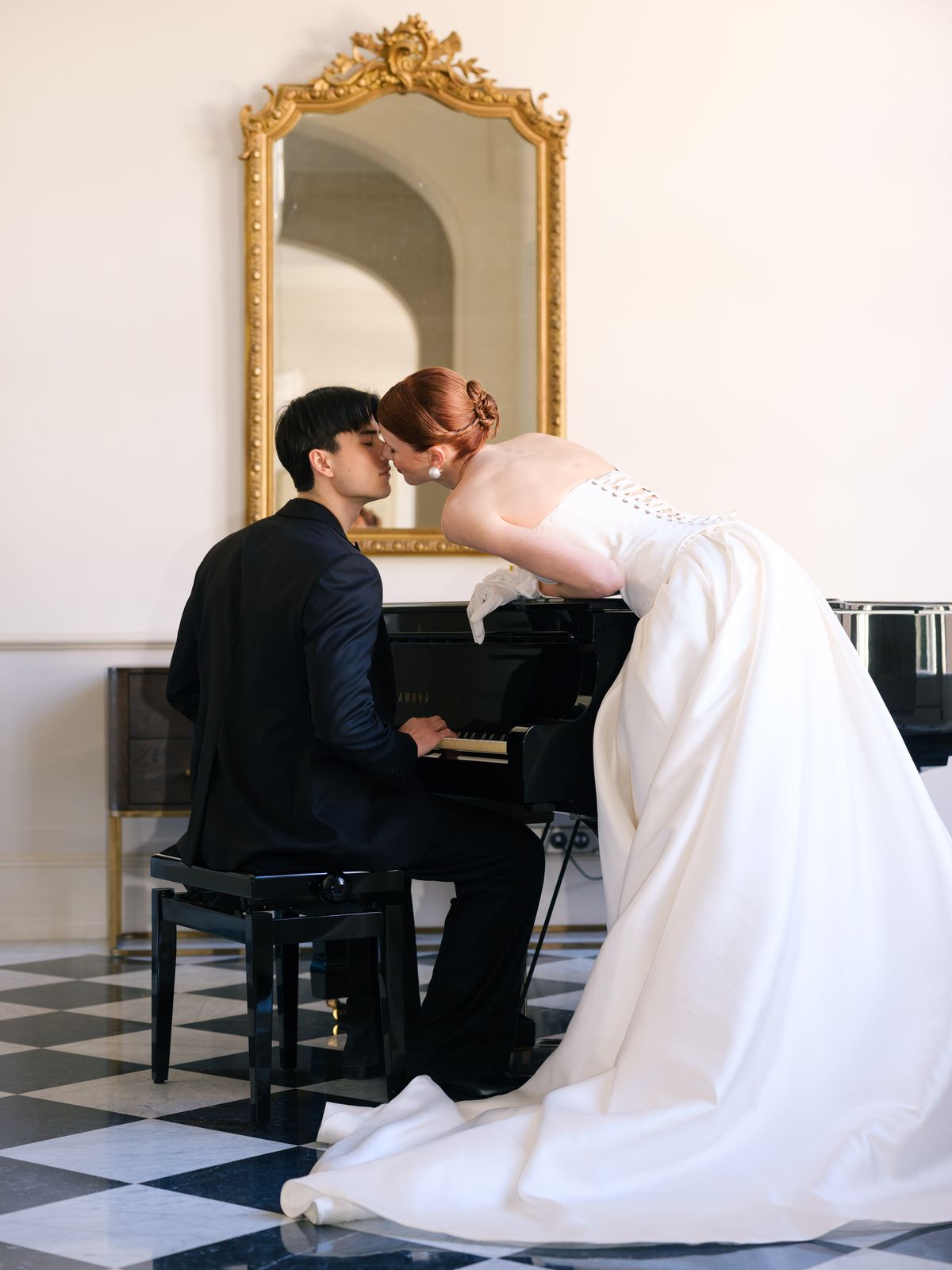 Bride leans across grand piano to kiss seated groom as satin gown spills across marble floor