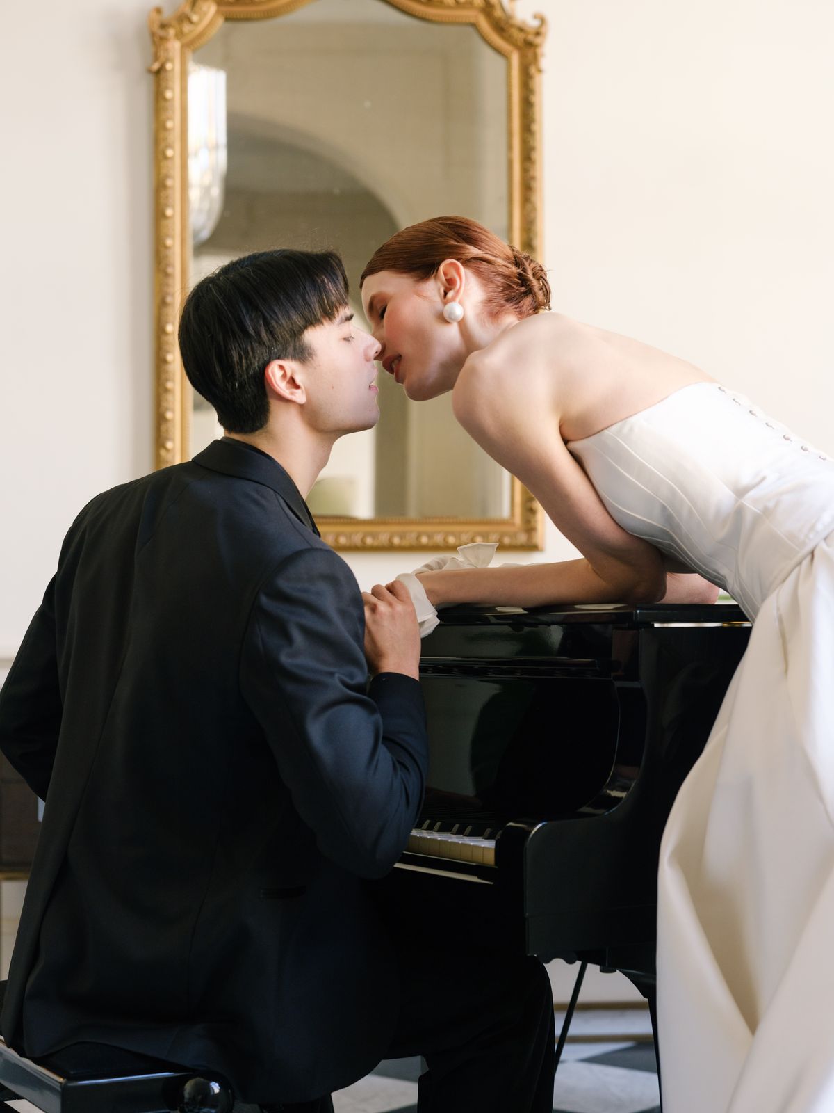 Bride and groom kiss across the open lid of a black grand piano beneath gilt mirror