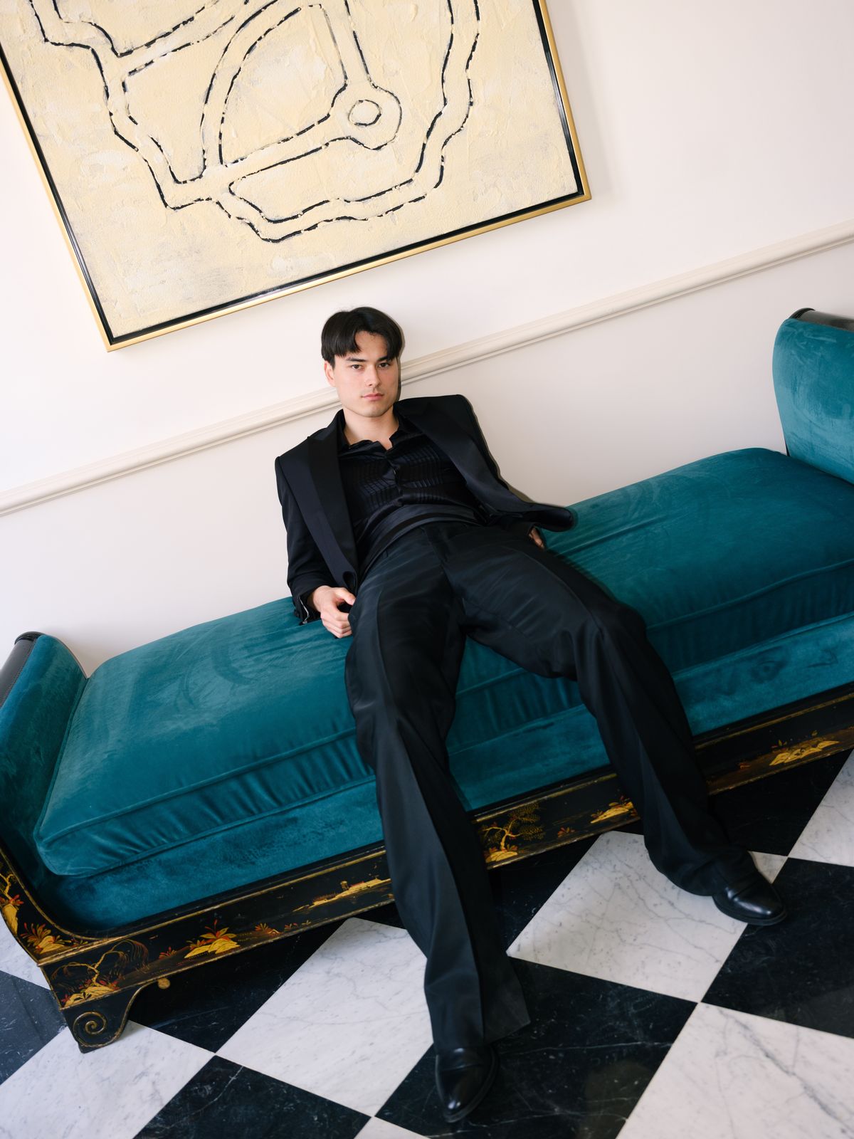Groom reclines across teal velvet daybed in black satin tuxedo beneath abstract canvas