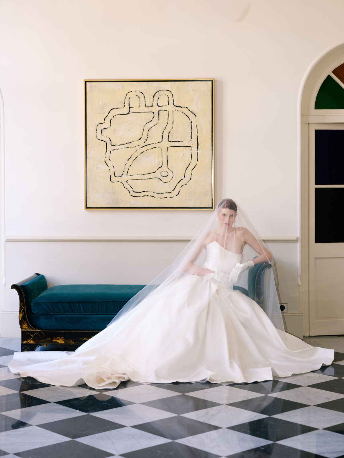 Bride sits on teal velvet daybed in voluminous satin A-line gown and cathedral veil with white gloves