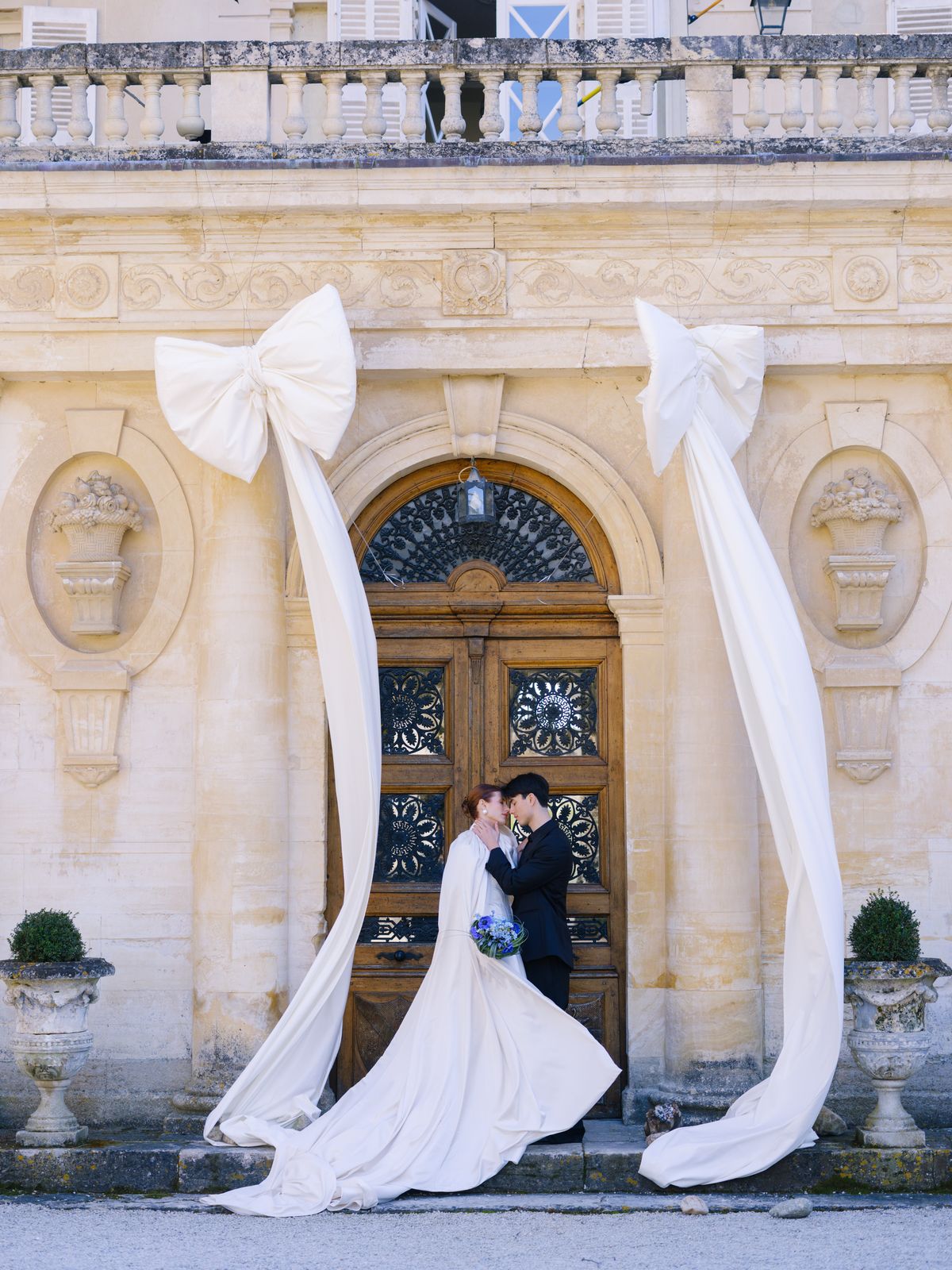 Couple kisses at arched château door framed by ivory satin bows with bride in long gown
