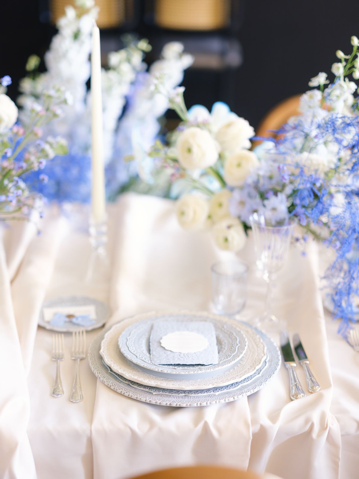 Place setting with lace chargers, blue menu and ranunculus delphinium centrepiece with ivory candle