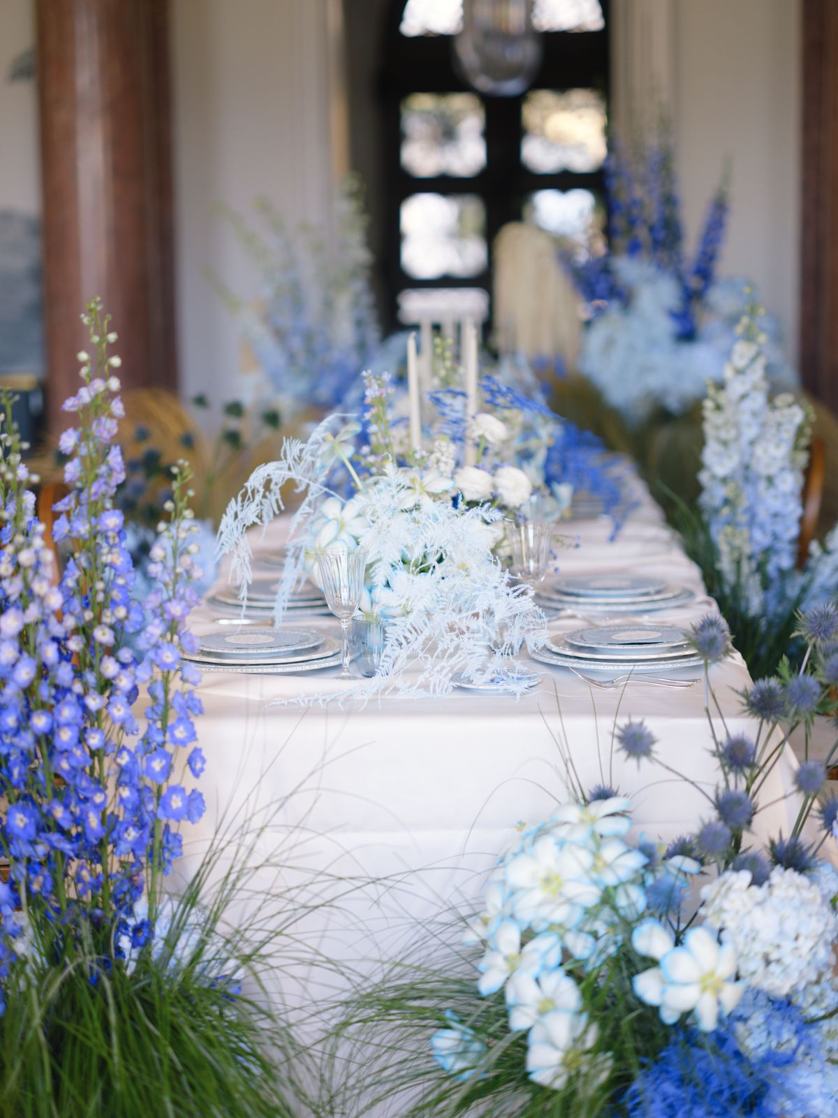 Long banquet table dressed with delphinium spires, eryngium thistle and silver chargers in château room