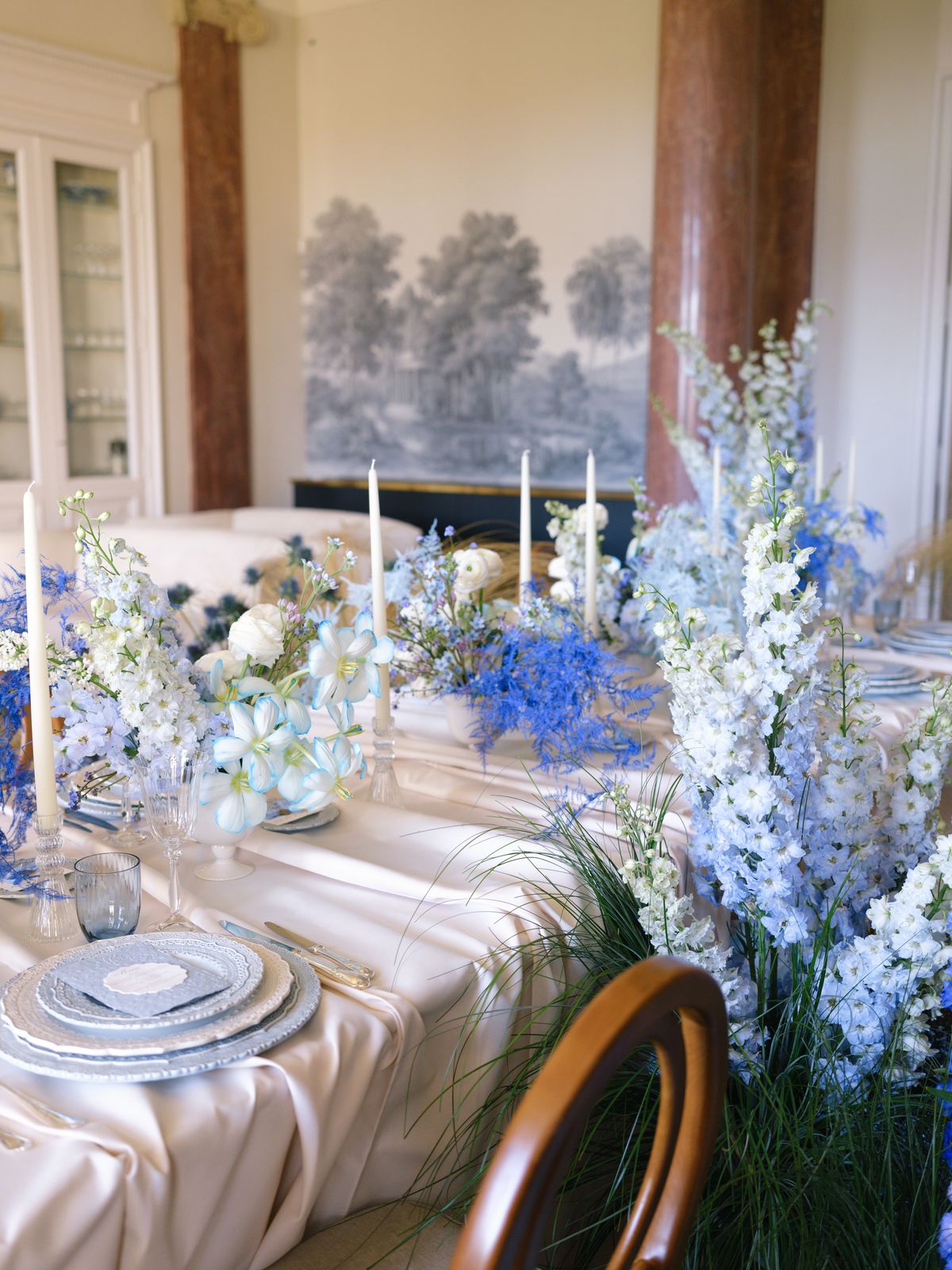 Angled banquet table with ivory tapers, delphinium and dried grasses beneath grisaille mural