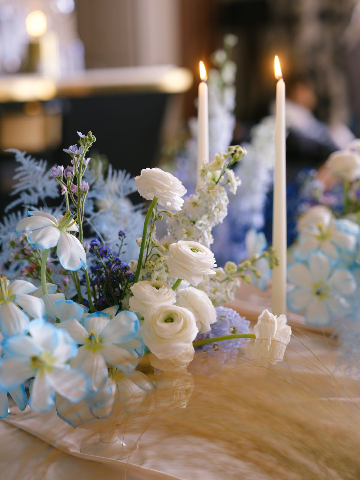 Close detail of white ranunculus, blue-tinted tulips and pampas grass beside lit ivory taper candles