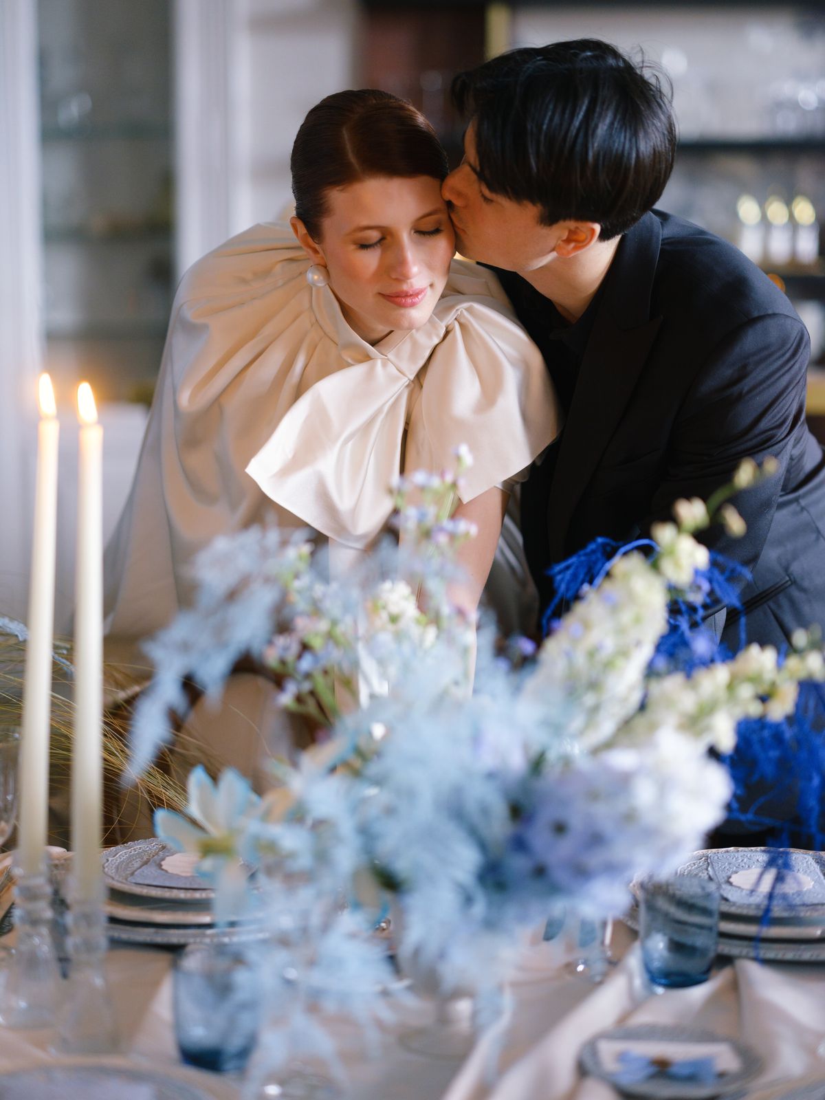 Groom kisses bride's temple as she wears dramatic ivory bow-collar cape over gown by candlelit table