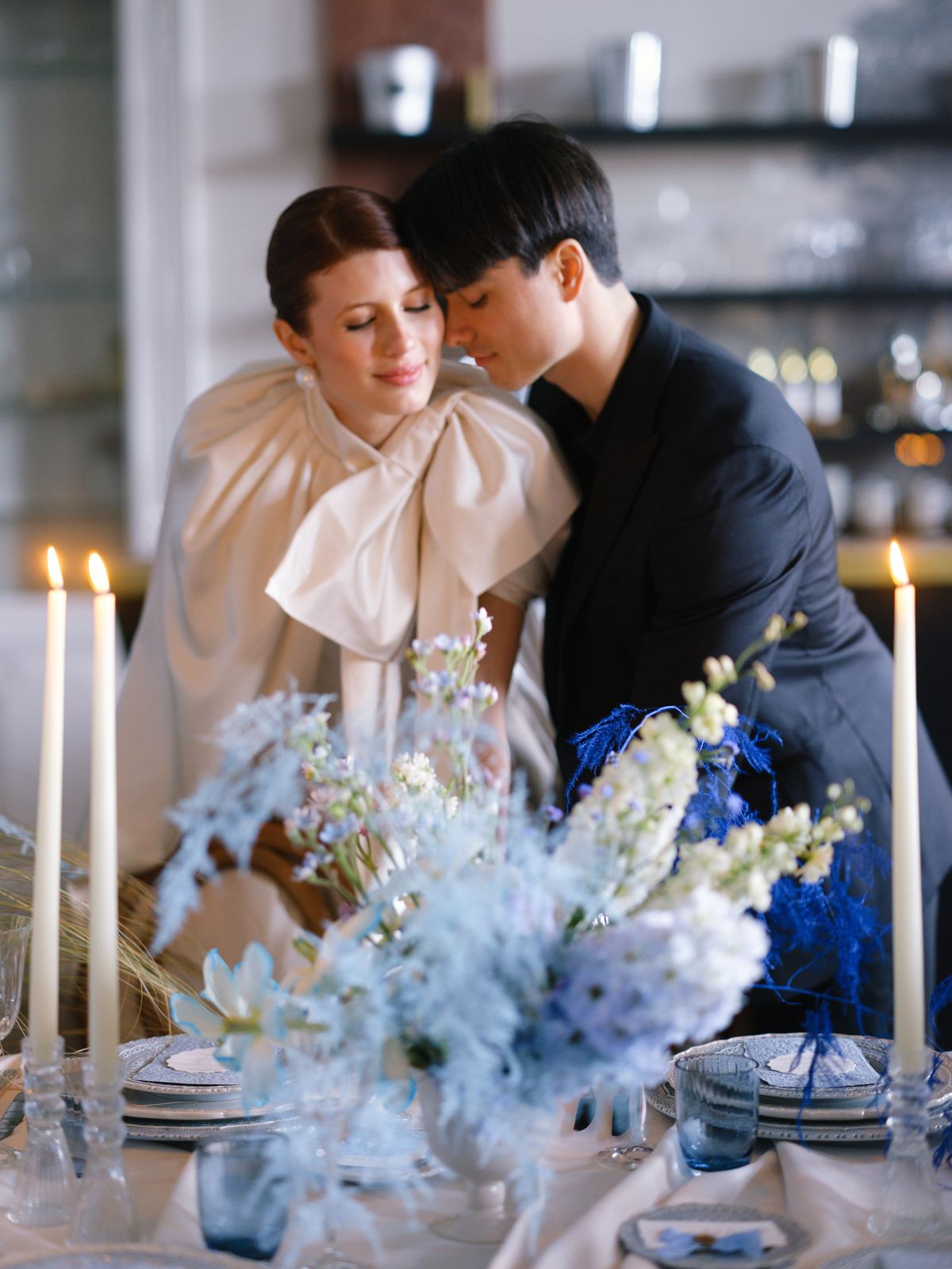 Couple cheek to cheek above candlelit table with bride in oversized ivory satin bow-collar cape