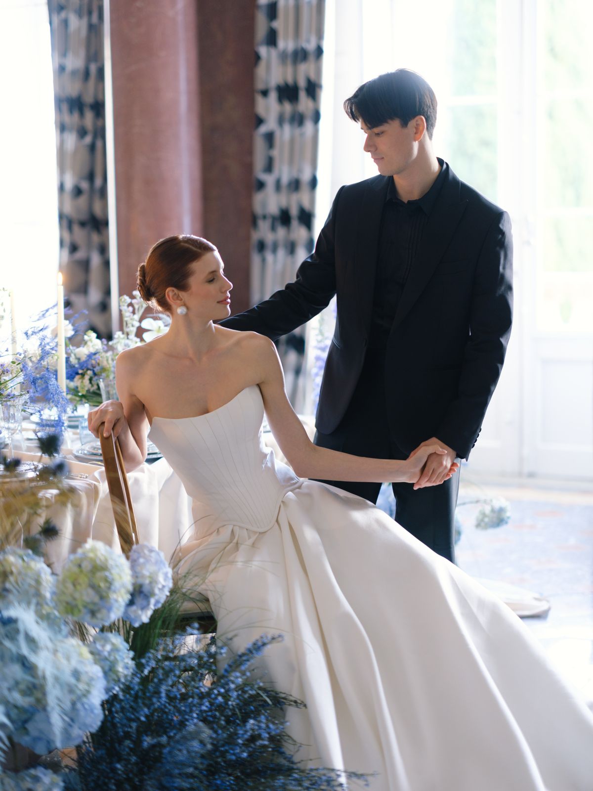 Groom helps bride rise from chair beside dressed banquet table with hydrangea and delphinium florals