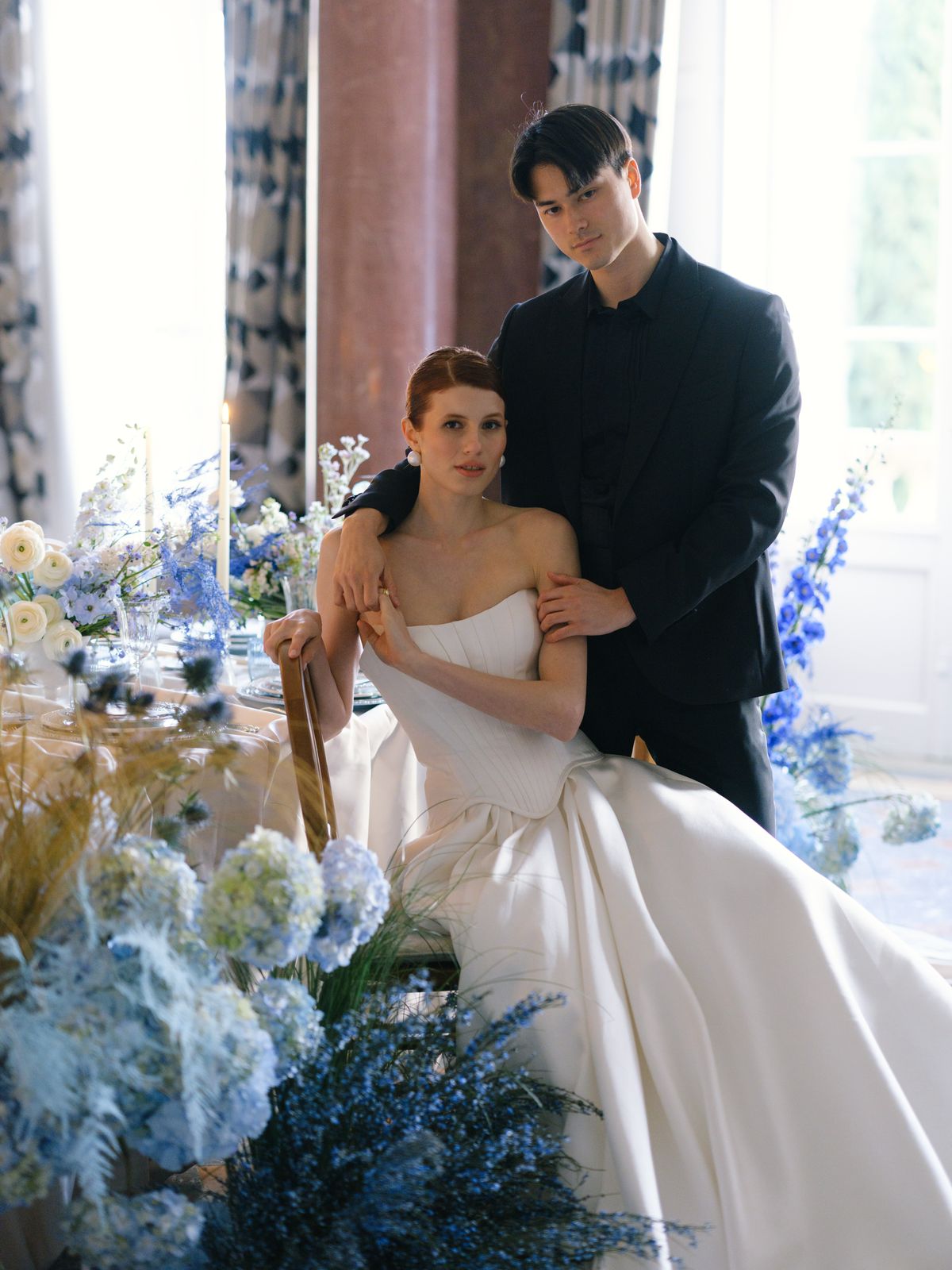 Bride seated in A-line satin gown with groom standing behind framed by blue hydrangea and delphinium