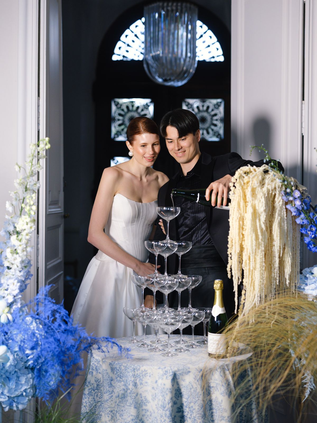 Couple pours champagne over coupe tower on toile-printed cloth beneath crystal chandelier