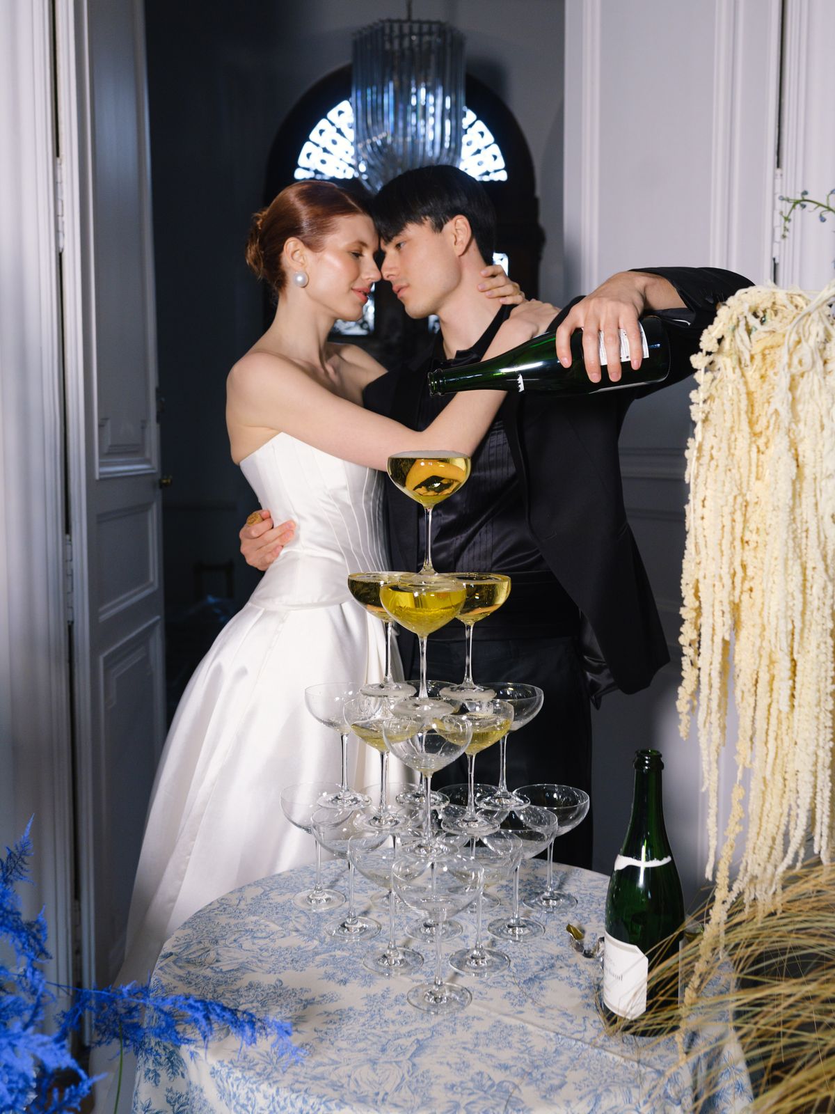 Couple embraces while pouring champagne over coupe tower with hanging amaranthus and blue florals