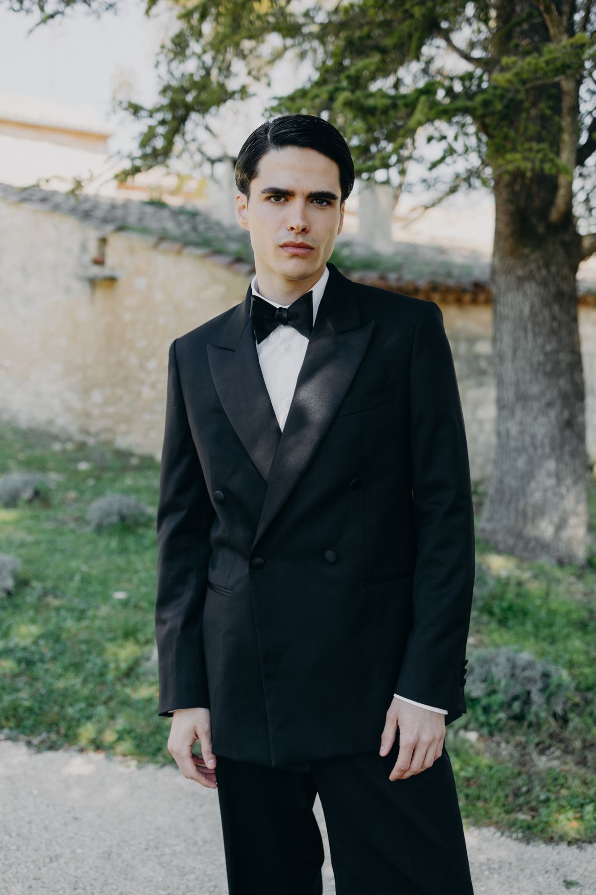Groom in a black tuxedo standing against the garden wall