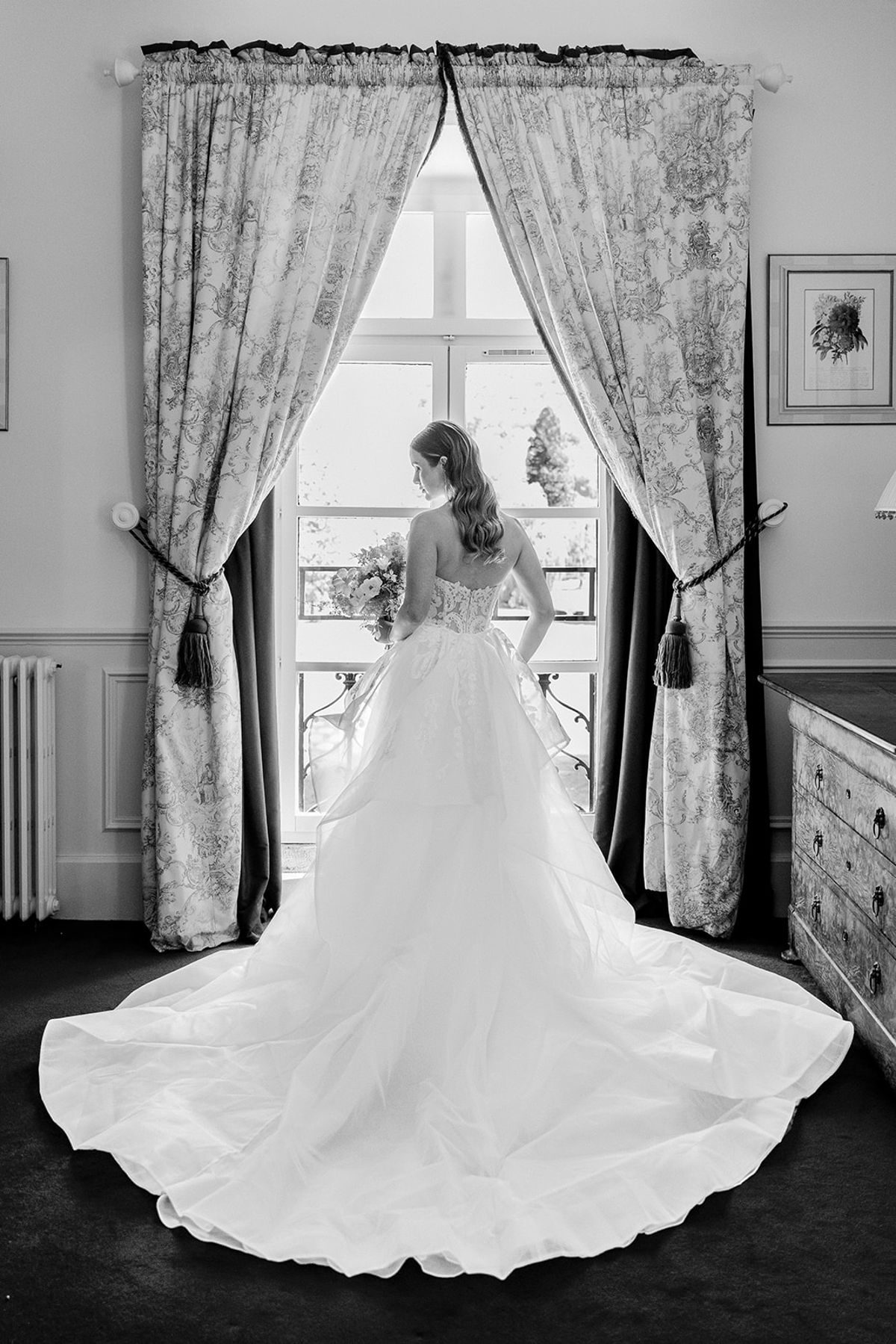 Black and white portrait of the bride at a window, the train of her gown spread behind her