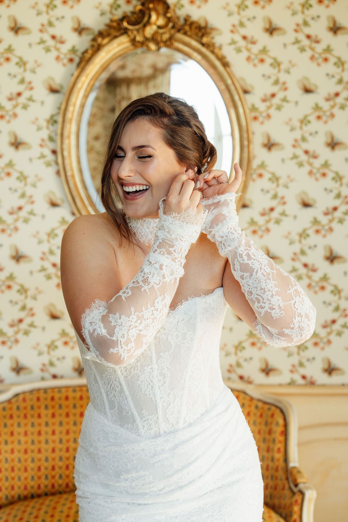Styled shoot models Harper and Liam: the bride laughing as she fastens an earring, Chateau de Mirambeau, spring editorial
