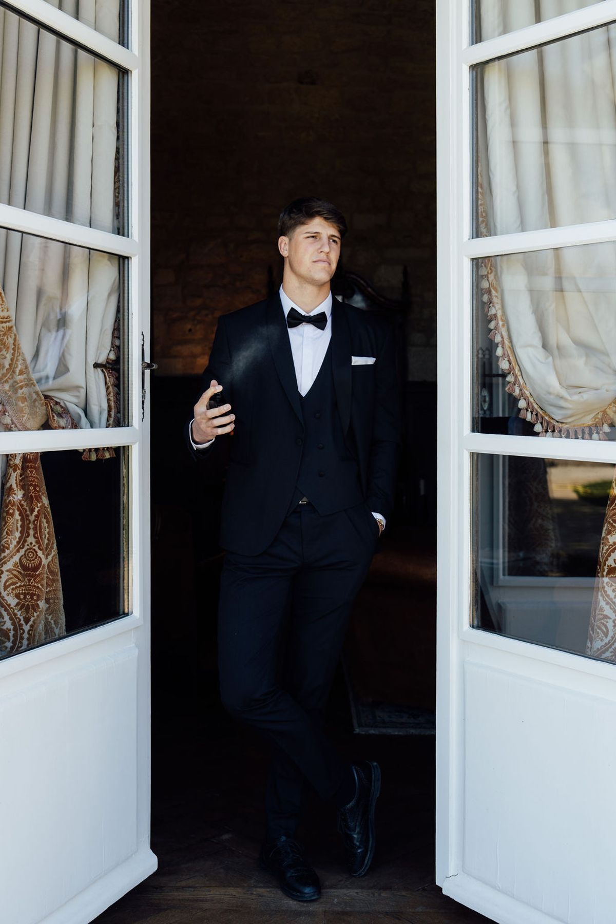 Styled shoot models Harper and Liam: the groom by a window adjusting his jacket, Chateau de Mirambeau, spring editorial