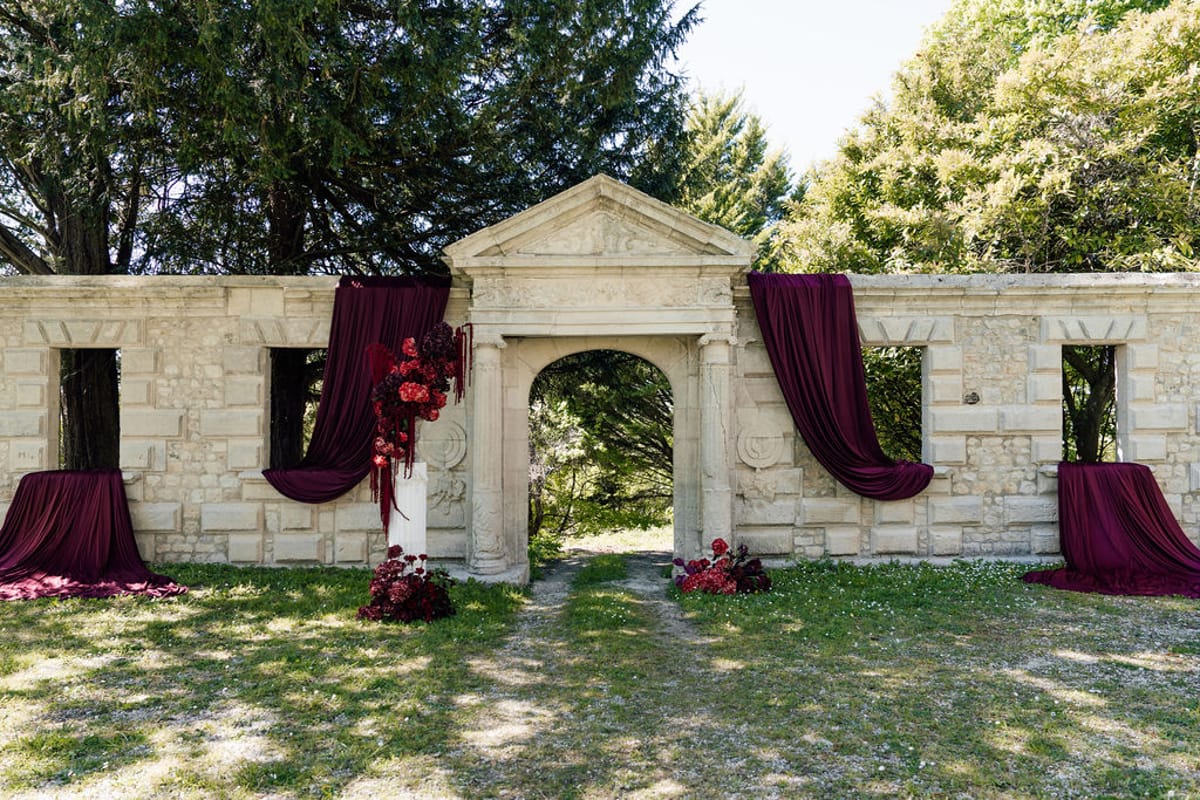 Styled shoot models Harper and Liam: the stone archway and burgundy drapery set for the ceremony, before the couple arrives, Chateau de Mirambeau, spring editorial