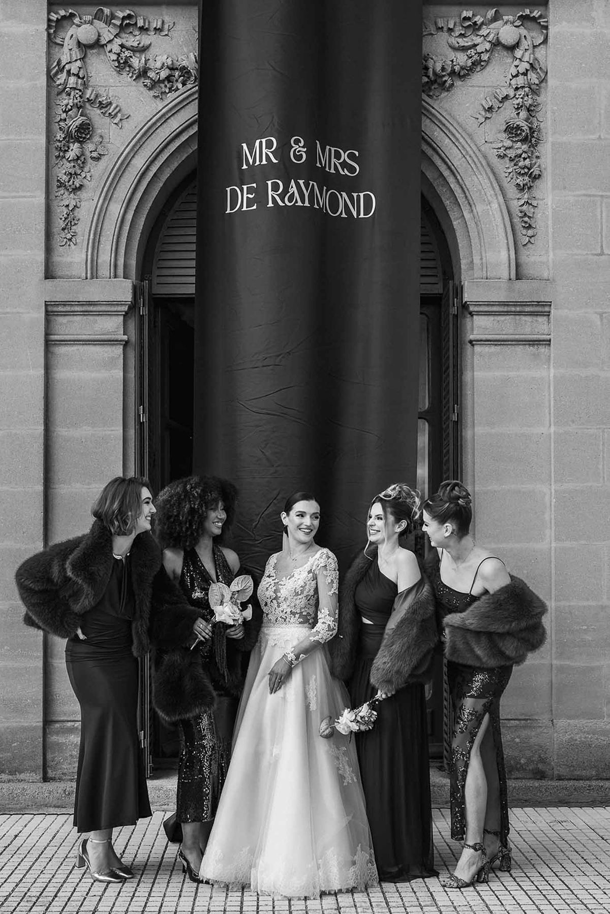Bride with bridesmaids under banner at Chateau du Terral entrance in black and white