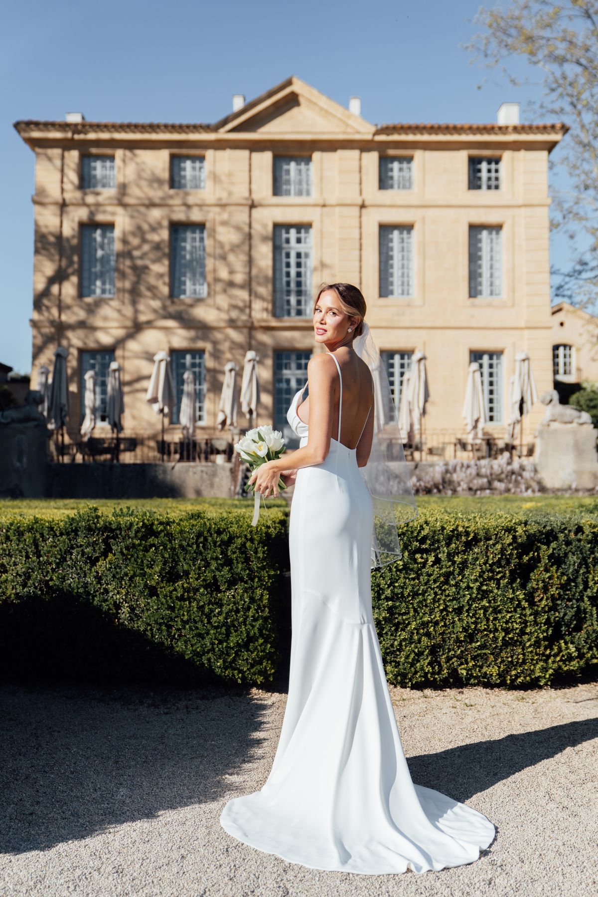Bride with a bouquet on the gravel walk before Chateau de la Gaude and its clipped hedges