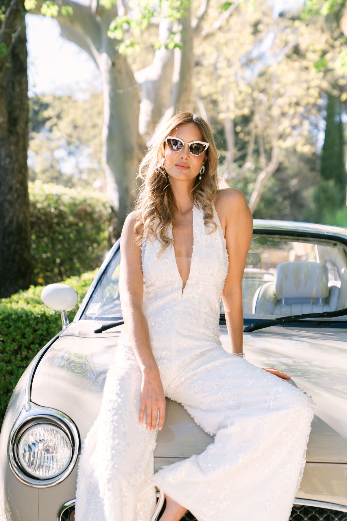 Bride in a sequinned jumpsuit and sunglasses seated on the bonnet of a vintage car