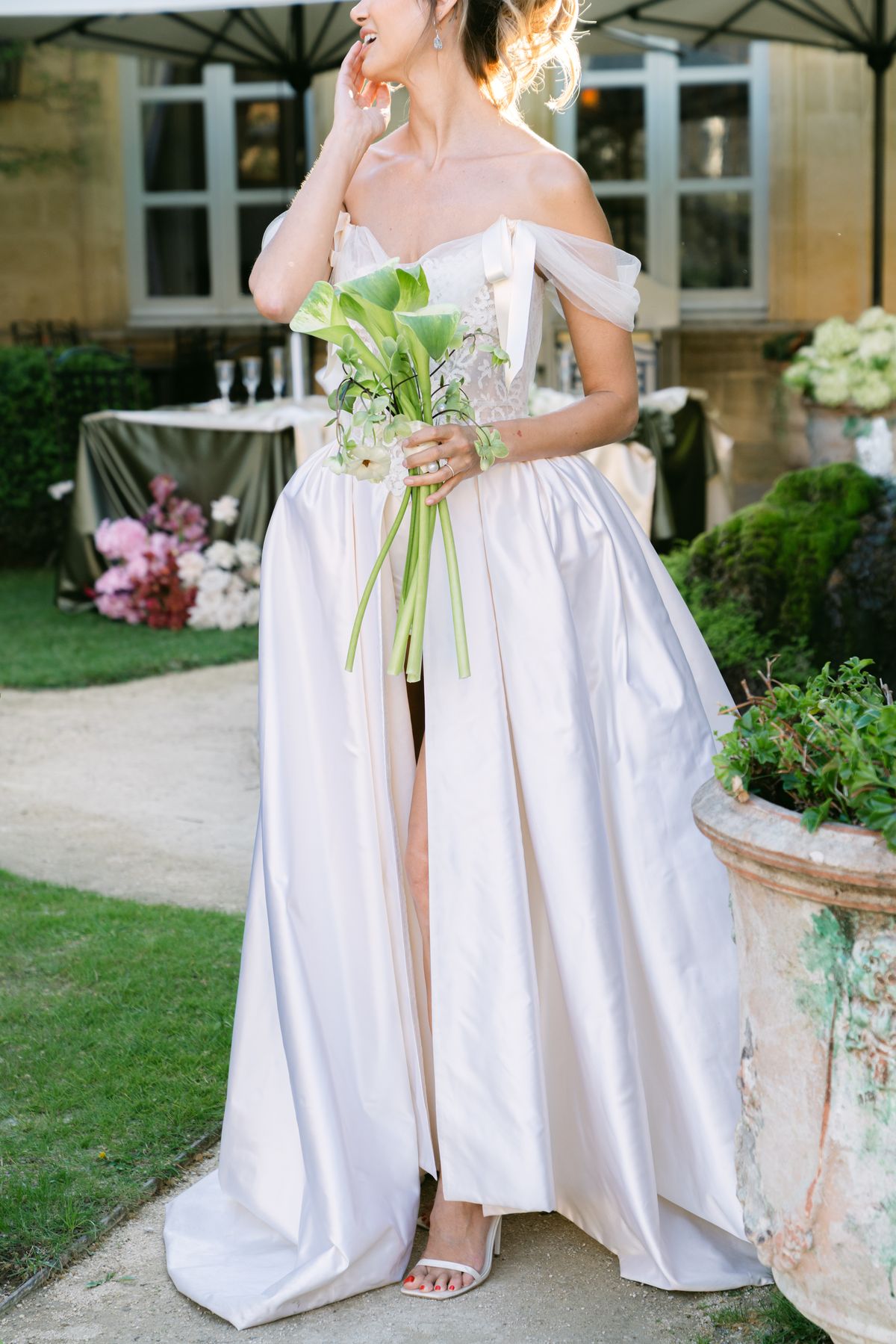 Bride in an off-shoulder gown with a thigh slit holding a calla lily bouquet on a lawn path