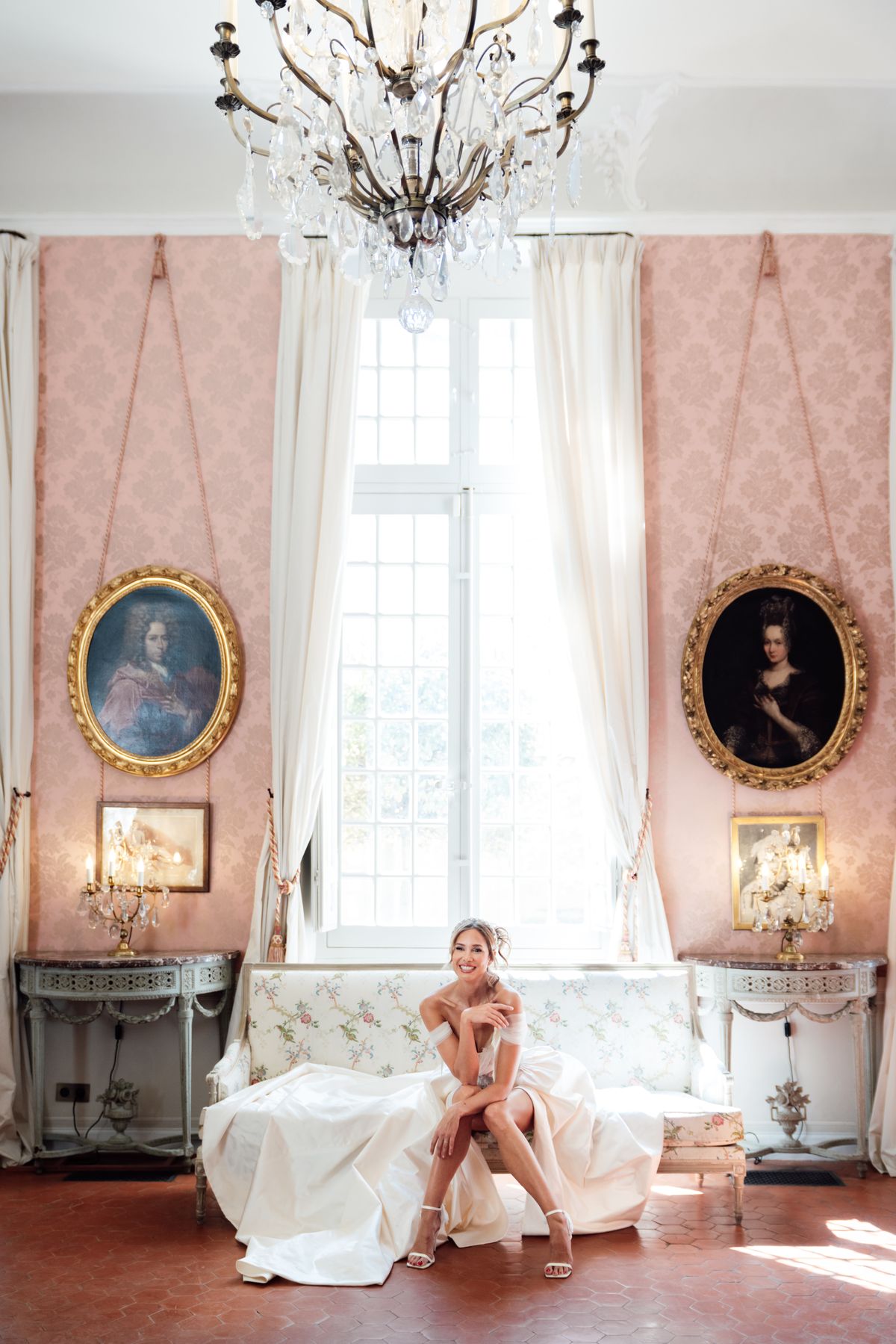 Bride seated in a rose-pink salon beneath a crystal chandelier with gilt-framed portraits behind