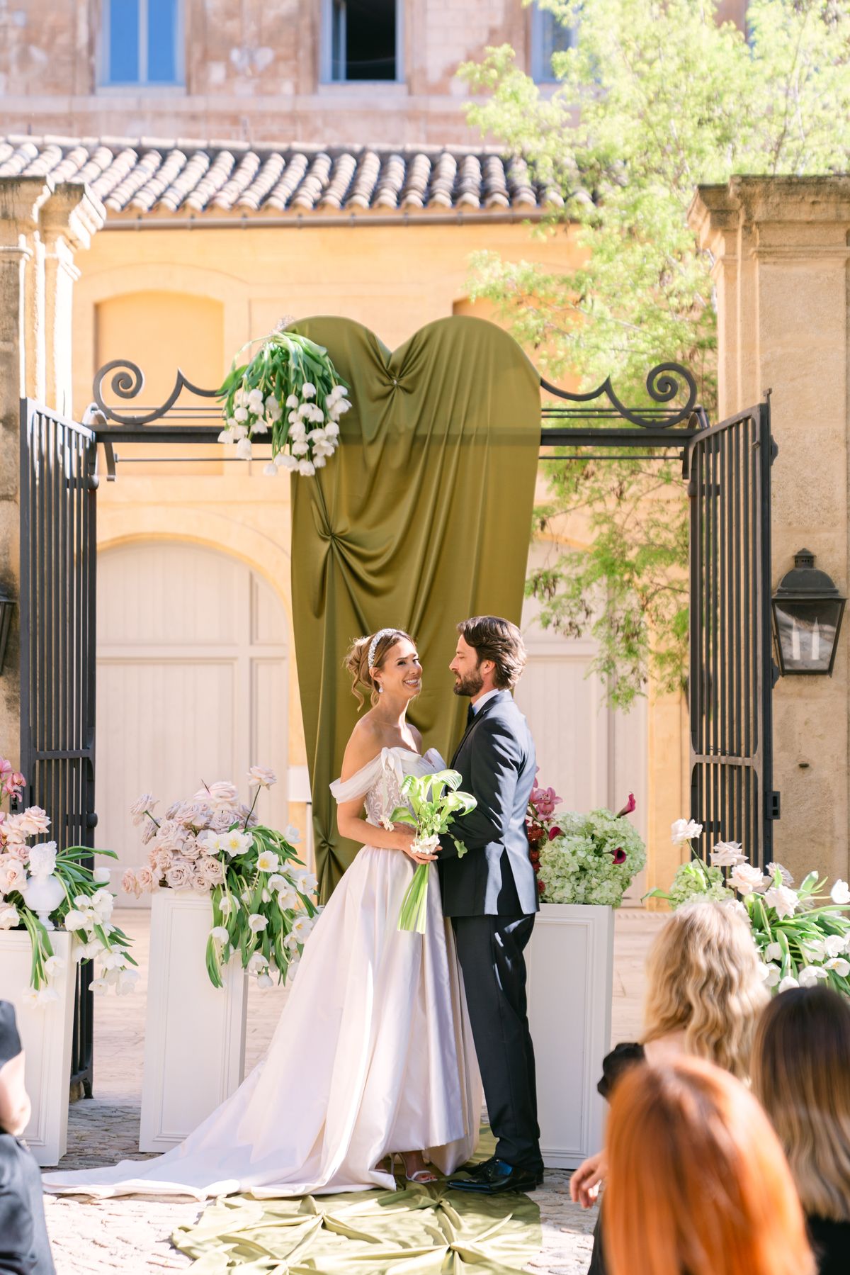 Couple standing face to face beneath an olive-green draped gateway during the ceremony