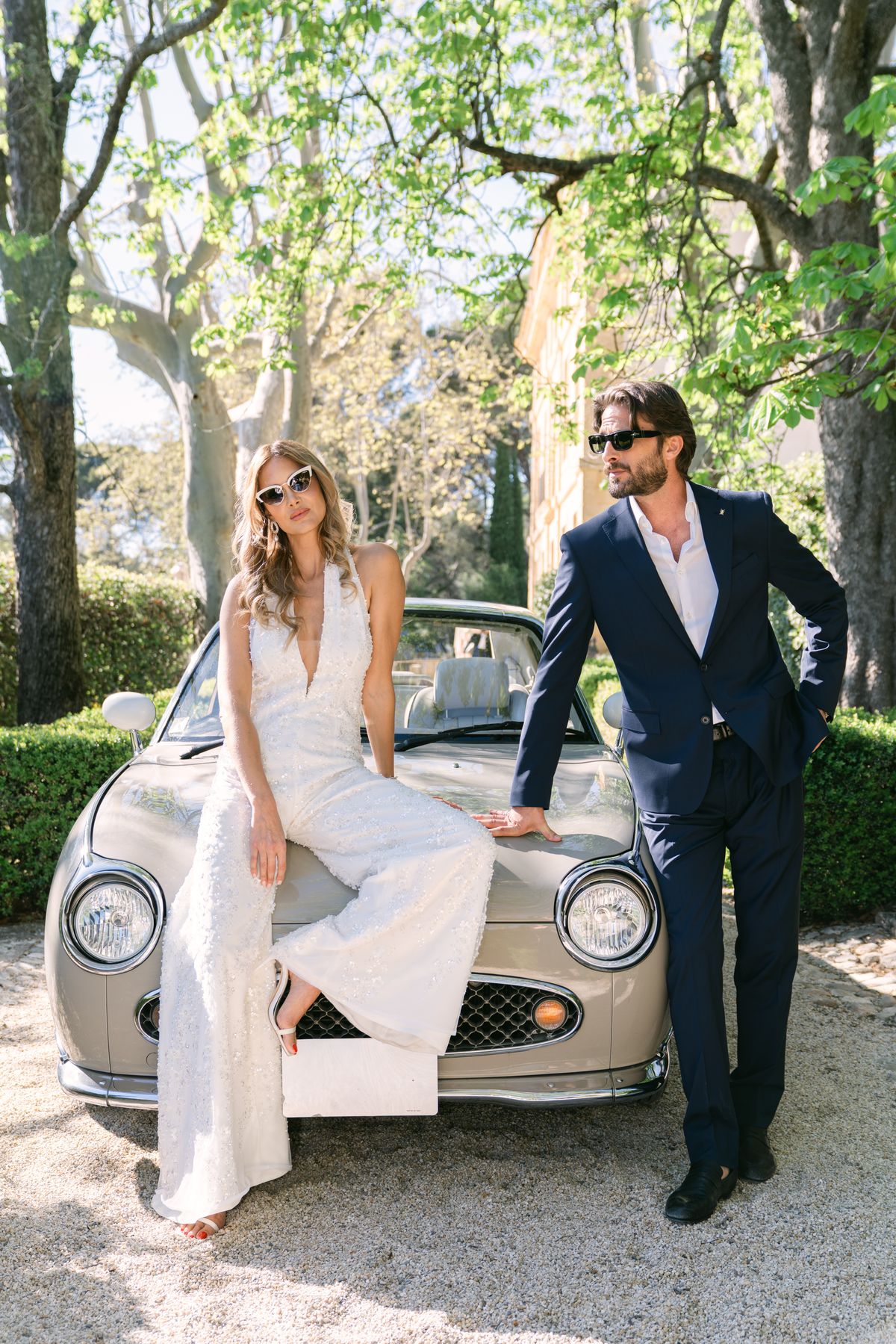 Couple in sunglasses beside a vintage car on a tree-lined drive