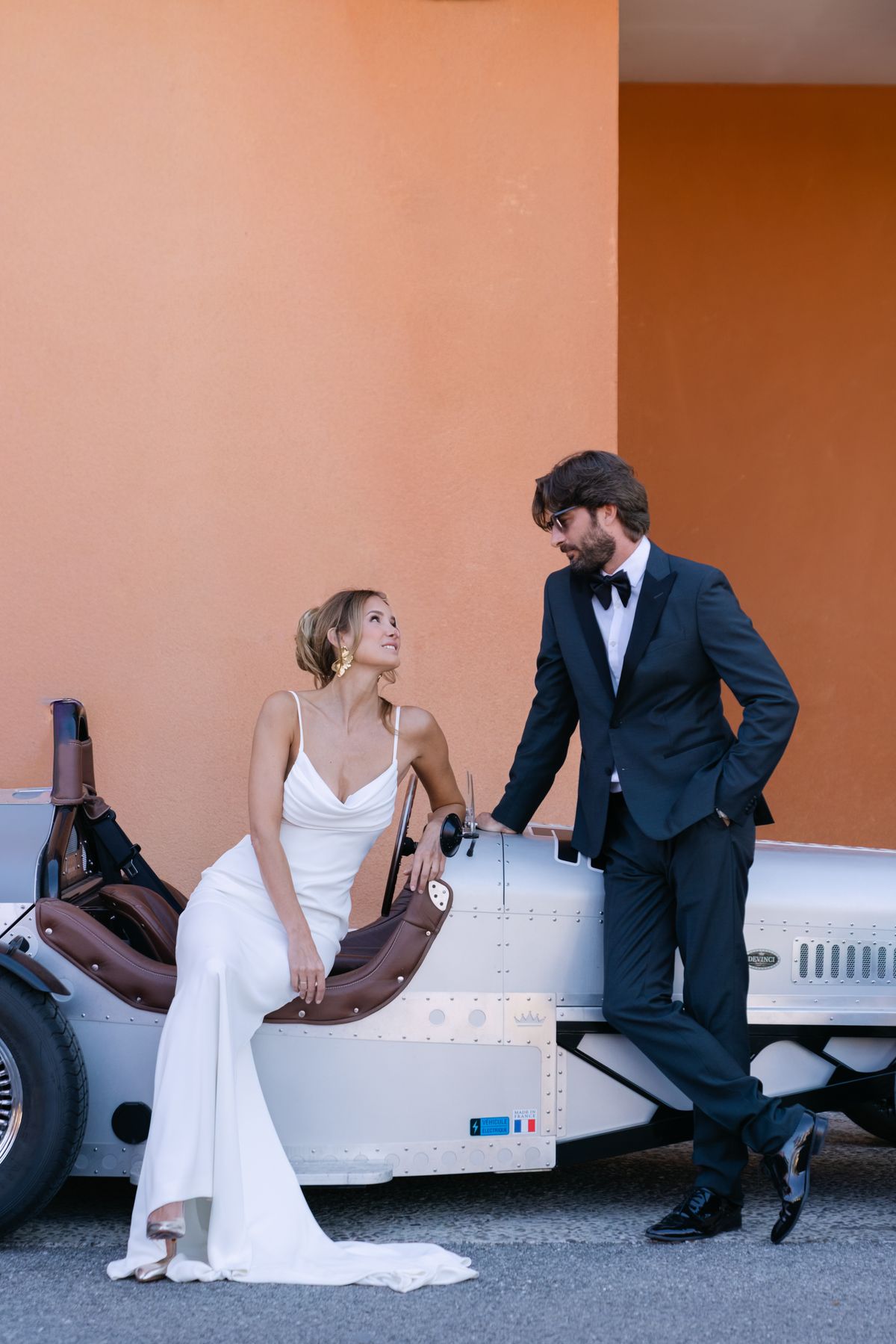 Bride seated on the bonnet of a vintage car with the groom standing beside her against a terracotta wall