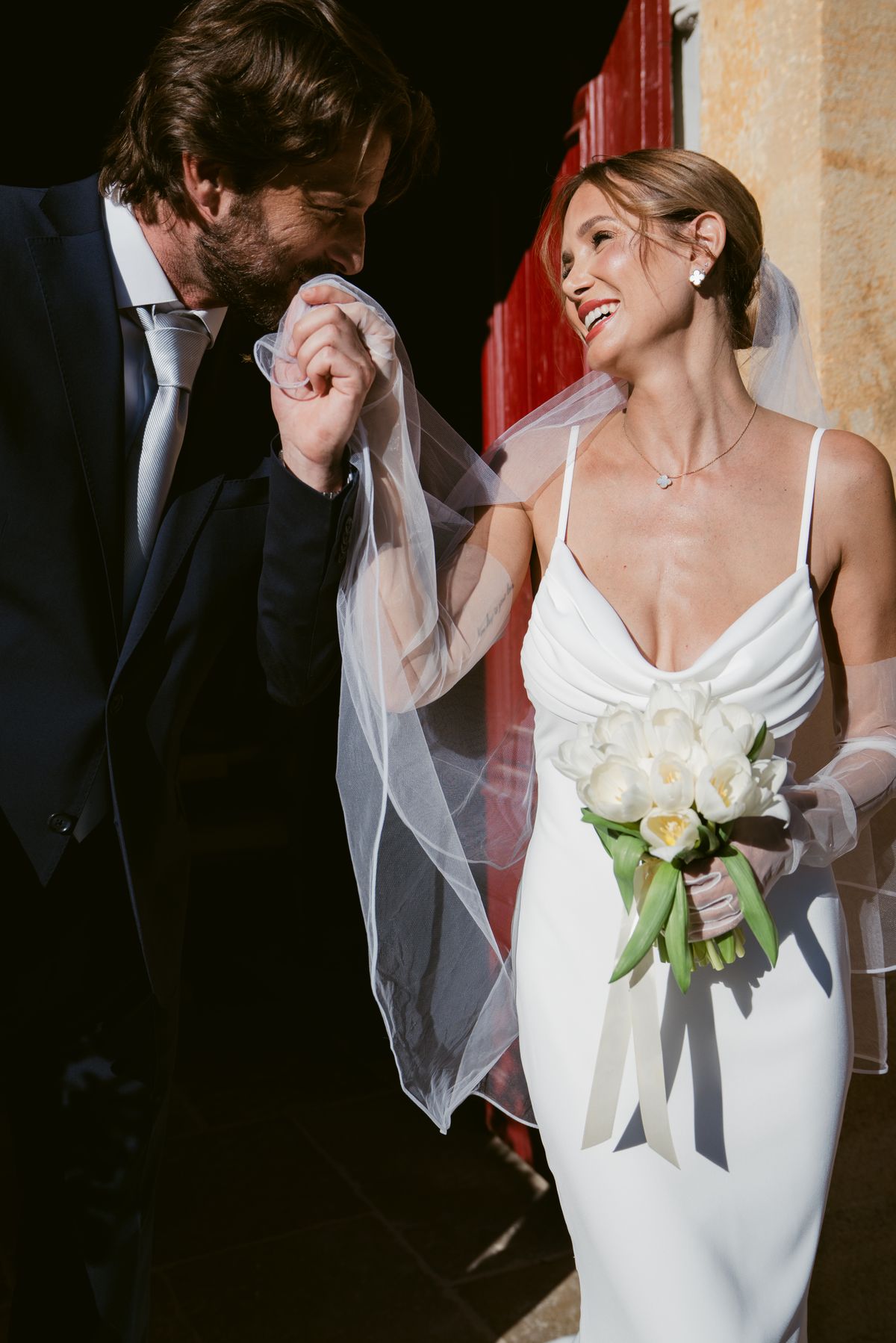 Groom kissing the bride’s hand beside a deep red door as she holds a tulip bouquet
