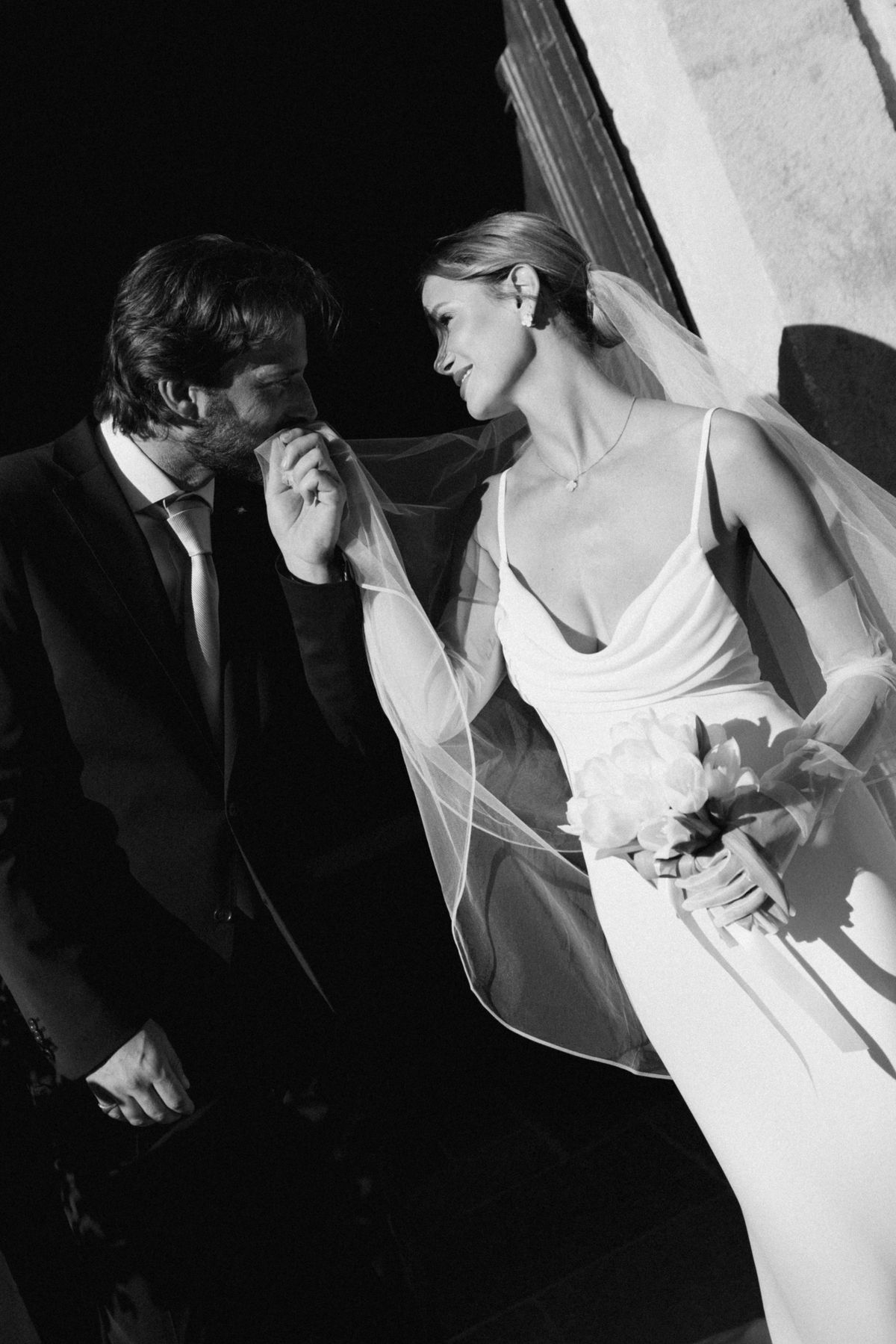 Black and white view of the groom kissing the bride’s hand with her veil lifted