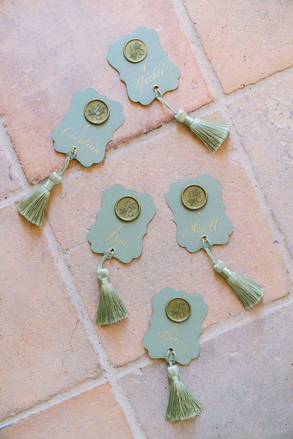 Five sage-green place cards with gold wax seals and tassels on terracotta tile