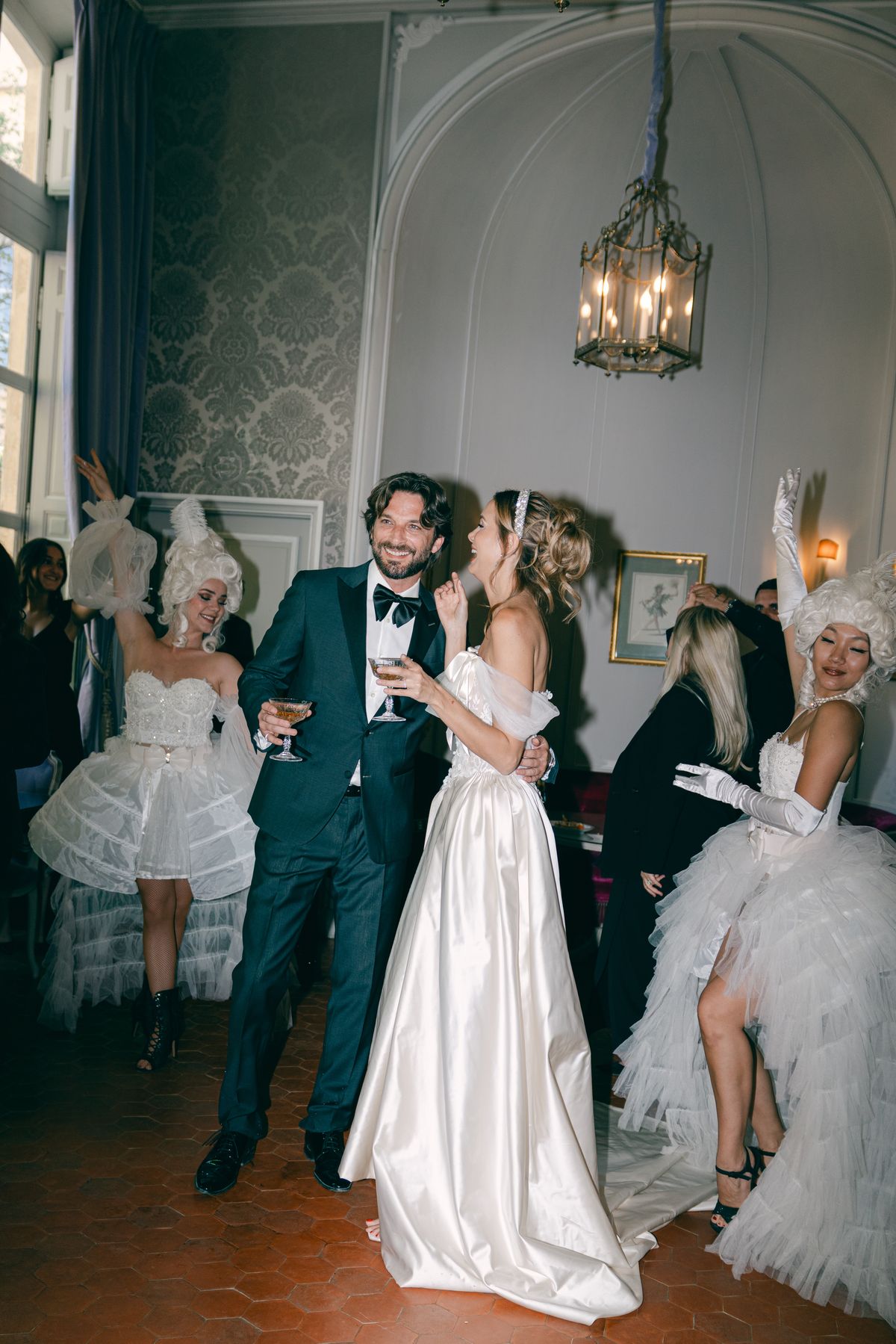 Couple holding champagne among costumed performers in a panelled salon